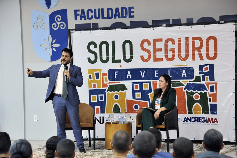 Foto horizontal da Abertura do Fórum Temático Solo Seguro Favela 2025. A foto mostra a palestra do juiz Hugo Abas. Ele está em pé, no lado esquerdo da imagem, falando em um microfone e gesticulando com a mão direita. Ele está vestindo um terno azul escuro e uma gravata. Sentada à direita dele, em uma cadeira, está uma mulher que está prestando atenção. Ela está vestindo um blazer escuro. Entre os dois, há uma pequena mesa redonda de madeira com garrafas de água. Ao fundo, há um banner grande que diz "SOLO SEGURO FAVELA" e, mais acima, a palavra "FACULDADE" e um logotipo azul. Na parte inferior do banner, são visíveis logotipos menores, incluindo um que diz "ANOREG/MA". Na parte inferior da foto, é possível ver as cabeças da audiência, indicando que há pessoas sentadas assistindo à apresentação.