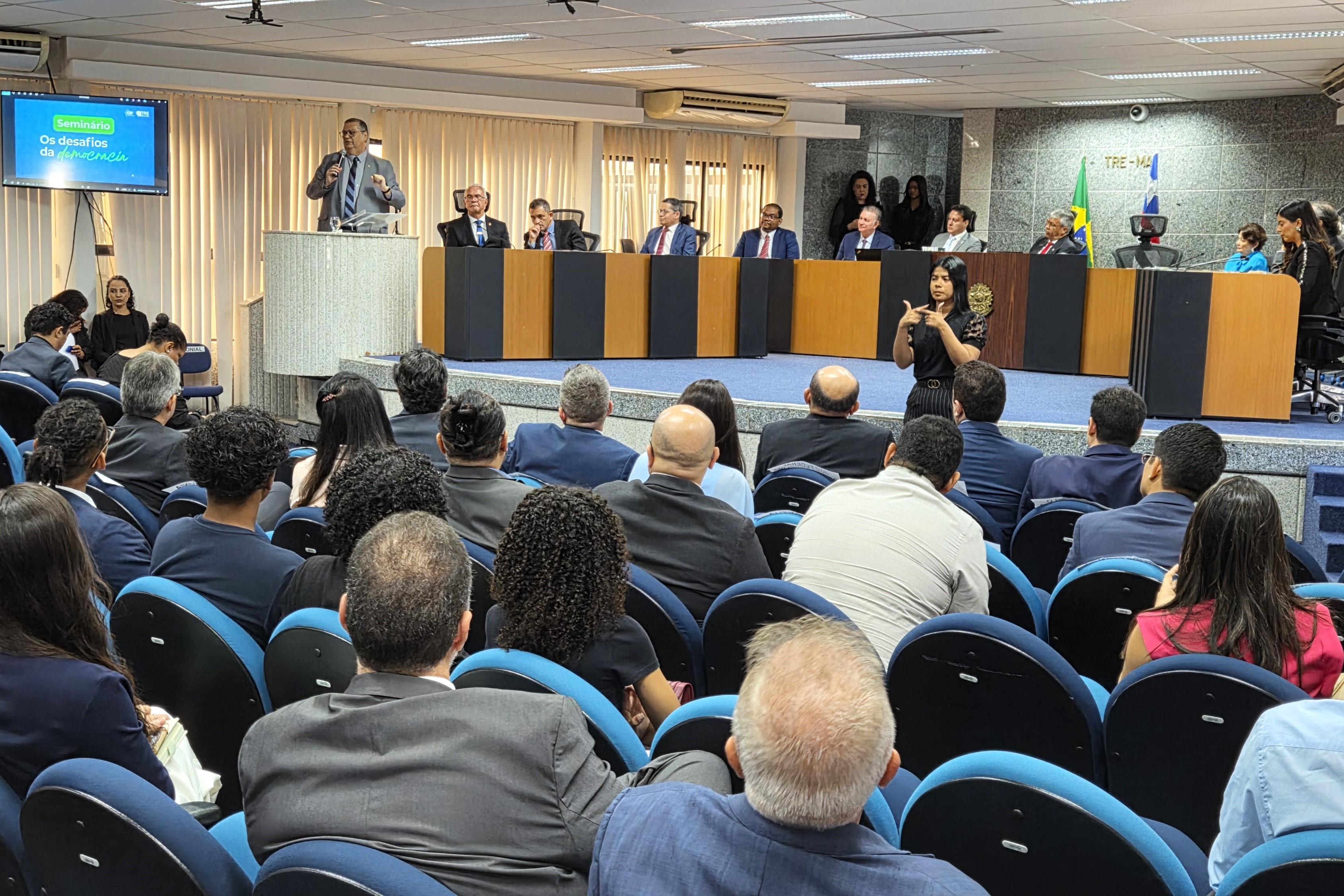 Presidente do TJMA participa de seminário sobre democracia e Justiça Eleitoral A imagem retrata um seminário com o tema "Os desafios da democracia", conforme aparece projetado na tela à esquerda. Um palestrante está falando em um púlpito, enquanto outras pessoas compõem a mesa de conferência ao fundo. A audiência assiste atenta, sentada em cadeiras azuis. À direita da imagem, uma intérprete de Libras traduz simultaneamente para a linguagem de sinais, promovendo acessibilidade ao evento. Também é possível ver a bandeira do Brasil ao fundo, indicando que o seminário ocorre em território nacional.