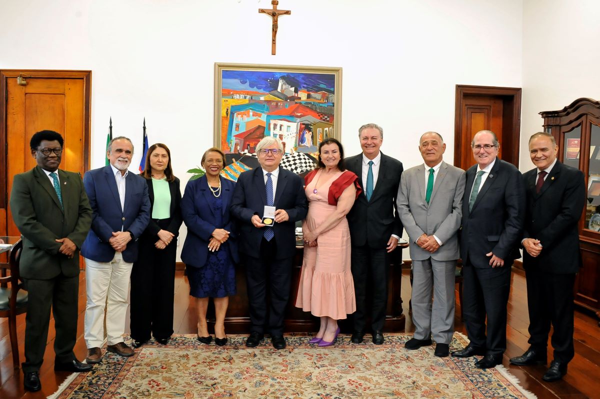 Foto horizontal de evento em que o ministro Herman Benjamin recebe medalha histórica em visita ao TJMA. Na foto, aparecem dez pessoas posando em um ambiente interno. No centro, o ministro, de terno, segura uma medalha dourada. Ao fundo, há uma cruz na parede, um quadro colorido e móveis de madeira. O ambiente é o gabinete da Presidência do TJMA.