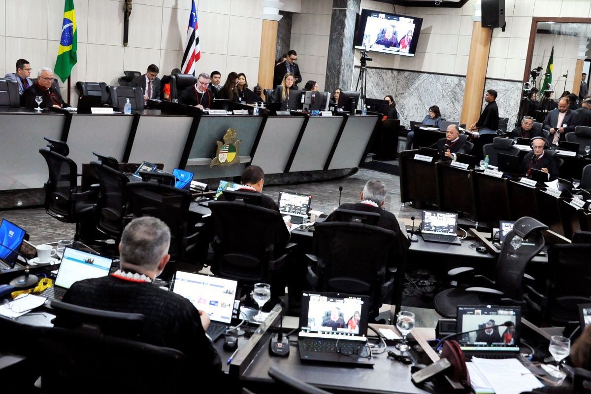 Foto horizontal da Sessão do Órgão Especial do TJMA em 4 de junho de 2025. A imagem mostra pessoas sentadas em bancadas, muitas delas usando computadores portáteis. Há um painel de vídeo grande ao fundo. Bandeiras, incluindo a do Brasil, são visíveis. O ambiente é a Sala das Sessões Plenárias do Tribunal, durante uma sessão.