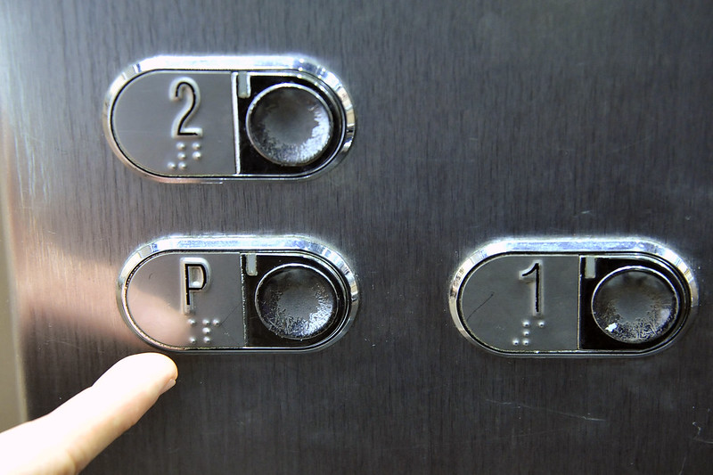 Foto horizontal mostra um painel de elevador, com três botões de metal para seleção de andar. No canto superior esquerdo, há um botão oval com o número "2" e a escrita em Braille para "2" logo abaixo. À direita, está a luz indicadora redonda. Abaixo do botão "2", no canto inferior esquerdo, há um botão similar com a letra "P" e a escrita em Braille para "P" logo abaixo. A luz indicadora redonda está à direita. No canto inferior direito, o terceiro botão exibe o número "1" com a escrita em Braille para "1" abaixo, e a luz indicadora redonda à direita. Um dedo indicador está pressionando o botão "P".
