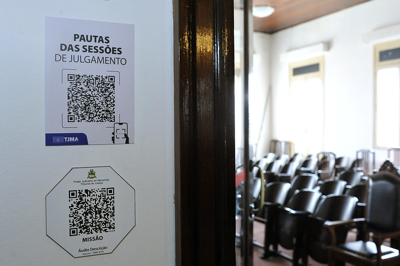 A foto mostra uma parede branca com dois cartazes fixados, ambos contendo códigos QR. O cartaz superior é retangular, com o texto "PAUTAS DAS SESSÕES DE JULGAMENTO" em letras grandes e pretas, acima de um código QR. Abaixo do código QR, há o logotipo do TJMA (Tribunal de Justiça do Maranhão) em azul e um ícone de celular com um código QR na tela. O cartaz inferior é octogonal, com o brasão do Poder Judiciário do Maranhão e o texto "Poder Judiciário do Maranhão Tribunal de Justiça" em sua parte superior. Abaixo, há outro código QR, e na parte inferior, o texto "MISSÃO" e "Áudio Descrição" com o logotipo do TJMA e "TJMA 2019". Entre os dois cartazes e ao lado deles, há uma moldura de porta escura, de madeira lustrosa. Ao fundo, através da porta, pode-se ver um salão com várias cadeiras alinhadas, algumas mais claras e outras mais escuras com estofamento em madeira, sugerindo um ambiente de audiência.