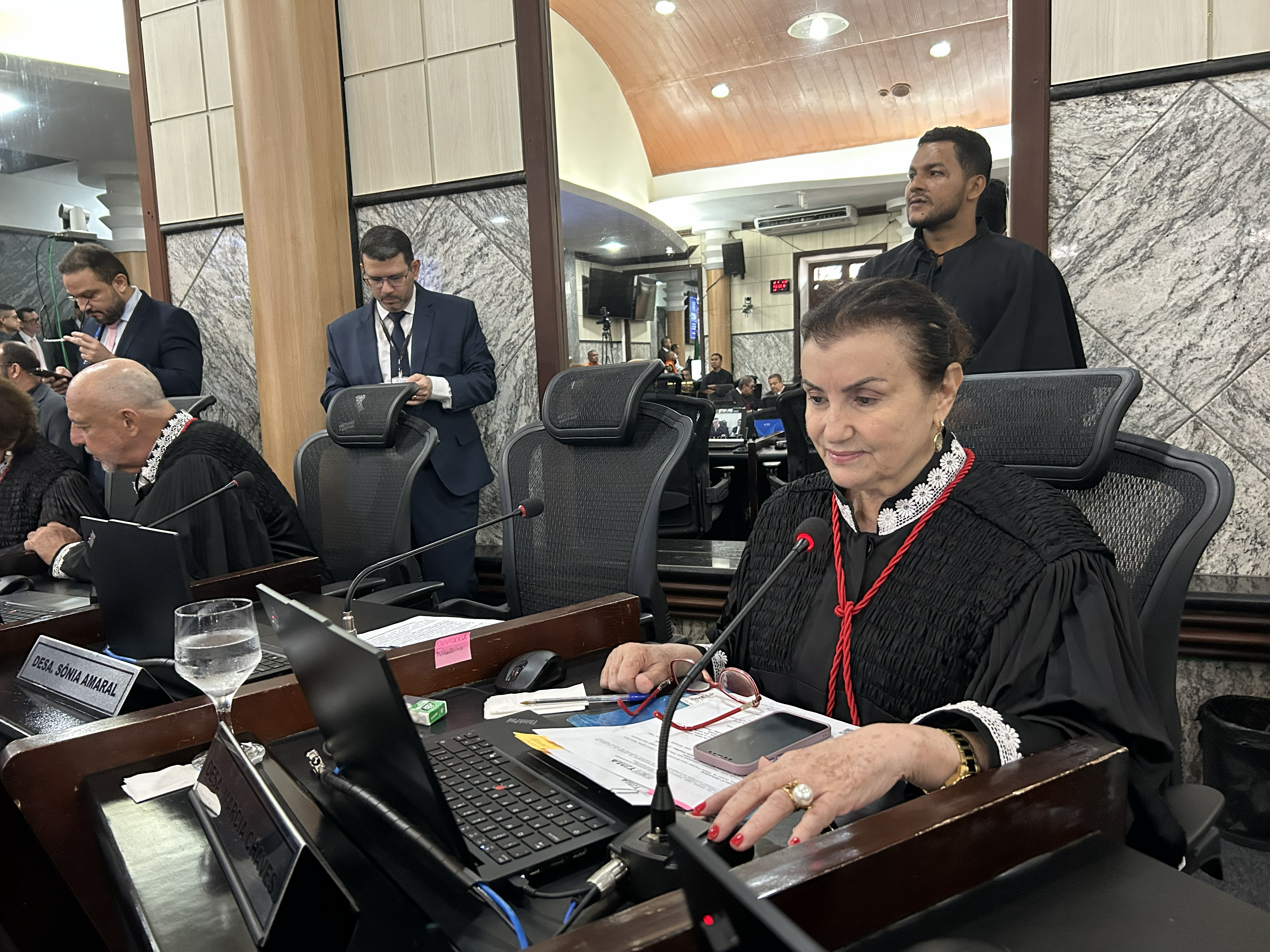 Em um ambiente semelhante, uma mulher vestindo toga preta com detalhes em renda e cordão vermelho sorri suavemente enquanto lê documentos à sua frente. Ao seu lado, outro juiz em toga preta lê em seu laptop. Ao fundo, homens em trajes formais conversam e observam a cena. Câmeras de vídeo estão posicionadas ao fundo para registrar a sessão.