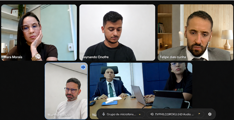 Foto horizontal NGF viabilização regularização e implantação de 50 unidades habitacionais em Senador Alexandre Costa. A imagem mostra a captura de tela de uma videoconferência com cinco quadrantes de participantes. Na fileira superior, há três janelas individuais identificadas. A fileira inferior apresenta um quadrante à esquerda e uma tela central maior com duas pessoas em uma sala de reuniões. Nessa sala, um homem de terno e gravata está sentado à mesa, de frente para a câmera, acompanhado por uma mulher ao seu lado que opera um notebook. A interface exibe ainda uma barra preta inferior com controles de áudio e configurações da plataforma de transmissão.