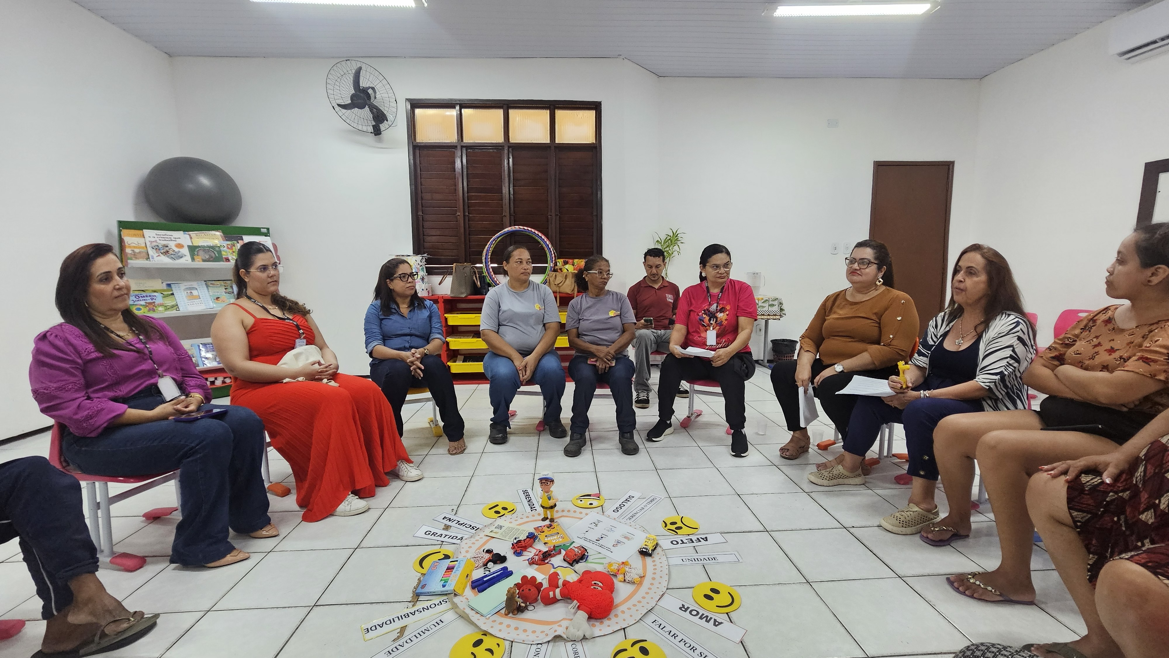 Em uma sala ampla e iluminada, onze pessoas estão dispostas em semicírculo sentadas em cadeiras e observam o centro do espaço. No chão de piso claro, há uma esteira circular decorada com placas com palavras como