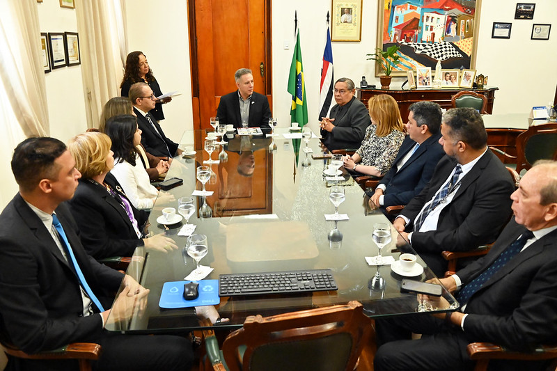 Em um escritório formal, cerca de doze pessoas estão reunidas em torno de uma grande mesa retangular de vidro. A maioria veste trajes sociais, como ternos e blazers, e estão sentadas em cadeiras de madeira estofadas, enquanto uma mulher em pé ao fundo segura papéis. Sobre a mesa reflexiva, há copos com água, xícaras de café, telefones celulares e um teclado de computador. Ao fundo, ao centro, destacam-se a bandeira do Brasil e a bandeira do estado do Maranhão entre portas e móveis de madeira. À direita, a parede é decorada com quadros, incluindo uma pintura colorida e fotografias porta-retratos.