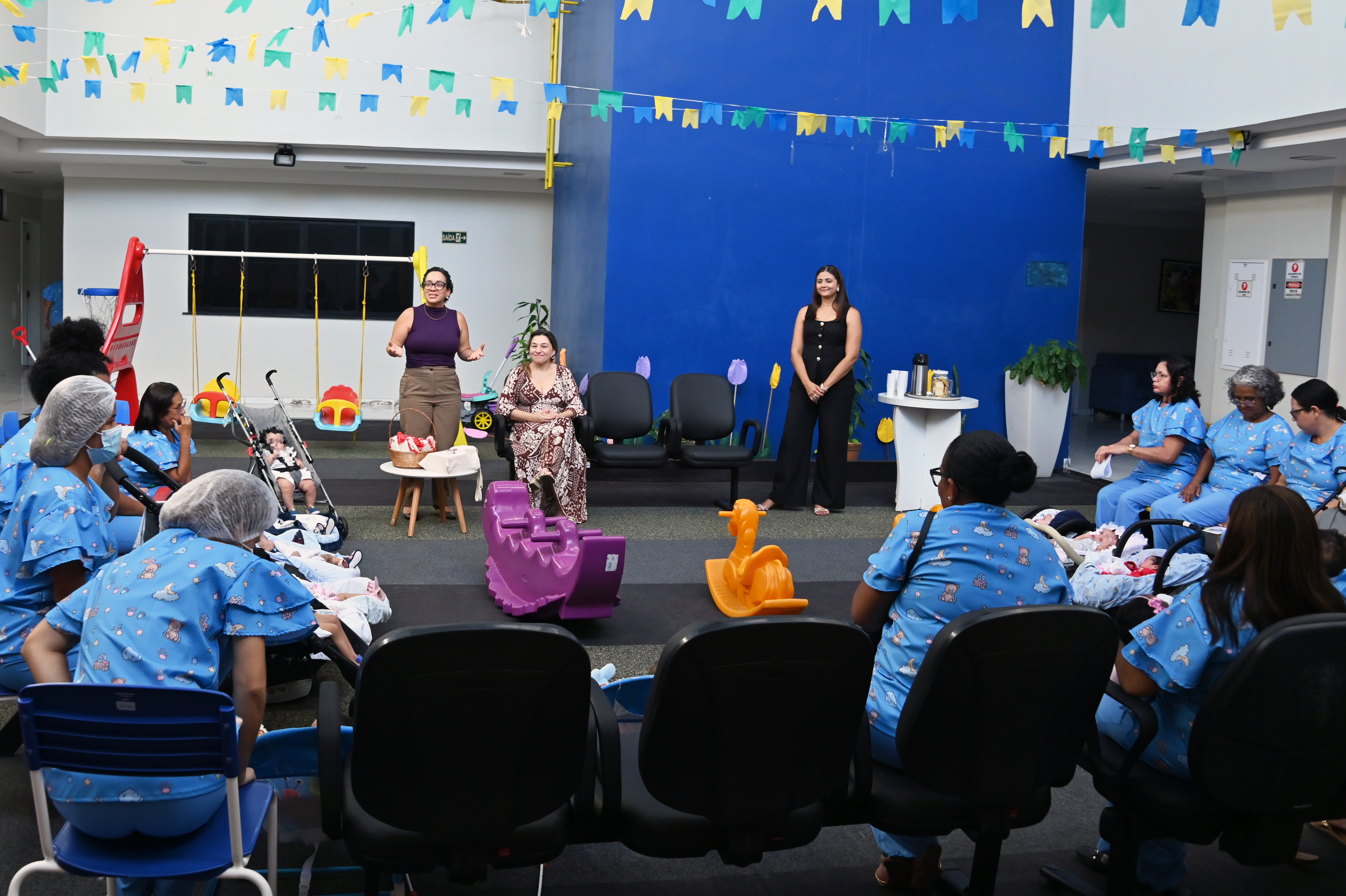 Um grupo de mulheres está reunido em círculo em um pátio interno decorado com bandeirinhas coloridas de festa junina. No centro, duas mulheres palestram: uma de blusa roxa em pé e outra de vestido estampado sentada. Elas são assistidas por várias outras mulheres vestindo uniformes azuis estampados, muitas delas com bebês no colo. Ao fundo, vê-se um balanço infantil, uma parede azul vibrante e mesas de apoio. Brinquedos de plástico em formato de cavalo e gangorra estão dispostos no chão de grama sintética.