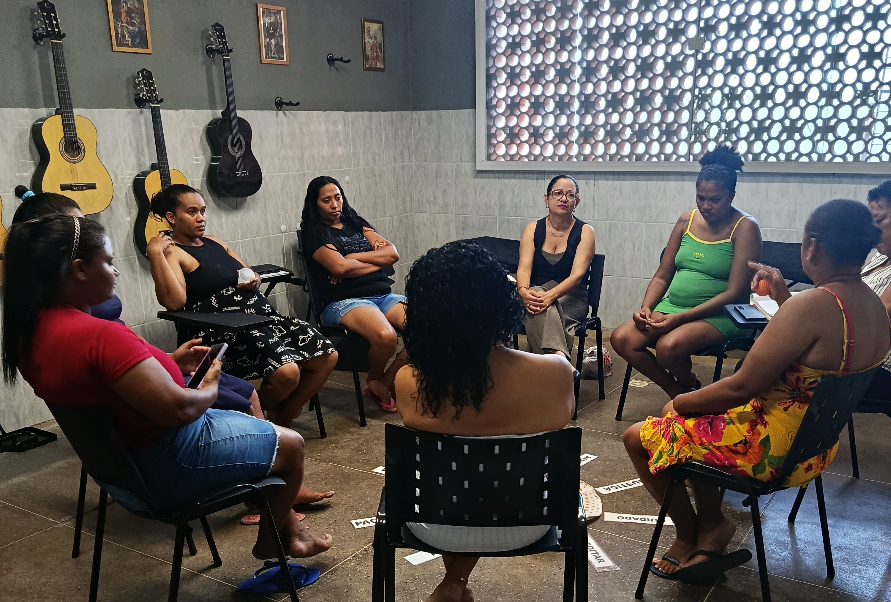 Um grupo de mulheres está sentado em círculo em cadeiras pretas com prancheta em uma sala acolhedora. No centro do chão, há placas de papel espalhadas com palavras como