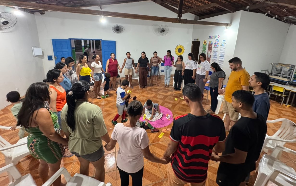 A imagem captura um momento de integração comunitária em um grande salão de eventos rústico. Cerca de vinte adultos estão dispostos em um grande círculo no centro da sala, de mãos dadas, todos voltados para o interior do círculo. No centro, duas crianças e uma adulta estão sentadas em um pano rosa estendido no chão, interagindo com objetos variados, enquanto o grupo as observa. A sala tem piso de lajotas estampadas, paredes brancas, portas e janelas azuis, e um teto de vigas de madeira expostas com ventiladores de parede. Cartazes com temas de
