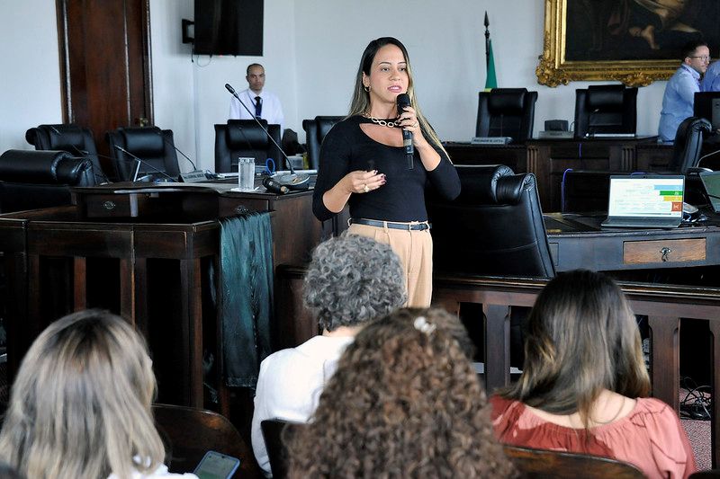 Foto horizontal da 2ª Reunião da RAE. Na imagem, há uma mulher de cabelo escuro e com blusa preta, calça bege e cinto, Bianca Soares, em pé, segurando um microfone enquanto fala para uma plateia. Ela está em uma sala com cadeiras e mesas de madeira escura, no Pleninho do TJMA. No fundo, há um homem em pé e, mais atrás, à direita, outro homem sentado. Em uma das mesas na frente, é possível ver um laptop aberto.