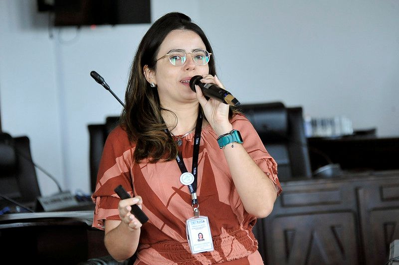 Foto horizontal da 2ª Reunião da RAE. Na imagem, há uma mulher de cabelo e olhos escuros, usando óculos de armação clara e um vestido com estampa em tons de rosa, Mariana Carneiro. Ela está segurando um microfone e um objeto preto na outra mão, enquanto fala. A mulher usa um crachá e um relógio com pulseira azul-petróleo. O fundo mostra um ambiente formal, com cadeiras escuras, mesas de madeira e equipamentos de áudio.