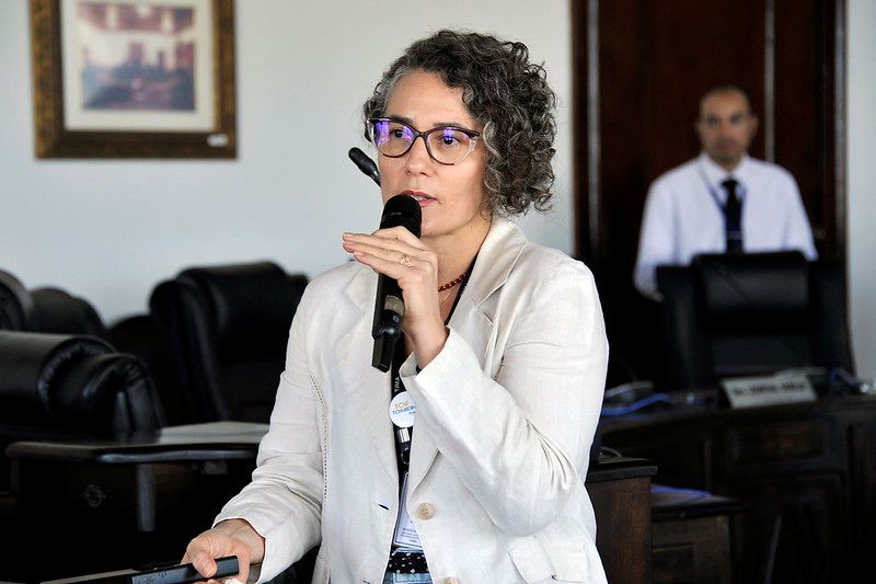 Foto horizontal da 2ª Reunião da RAE. Na imagem, uma mulher com cabelo cacheado e grisalho, usando óculos de armação transparente, Vanessa Gomes, que está em pé e segurando um microfone. Ela veste um blazer claro sobre uma blusa estampada. No fundo, é possível ver um homem de camisa branca e gravata escura, além de móveis de madeira e cadeiras de couro pretas, no Pleninho do TJMA. Há também um quadro pendurado na parede à esquerda.