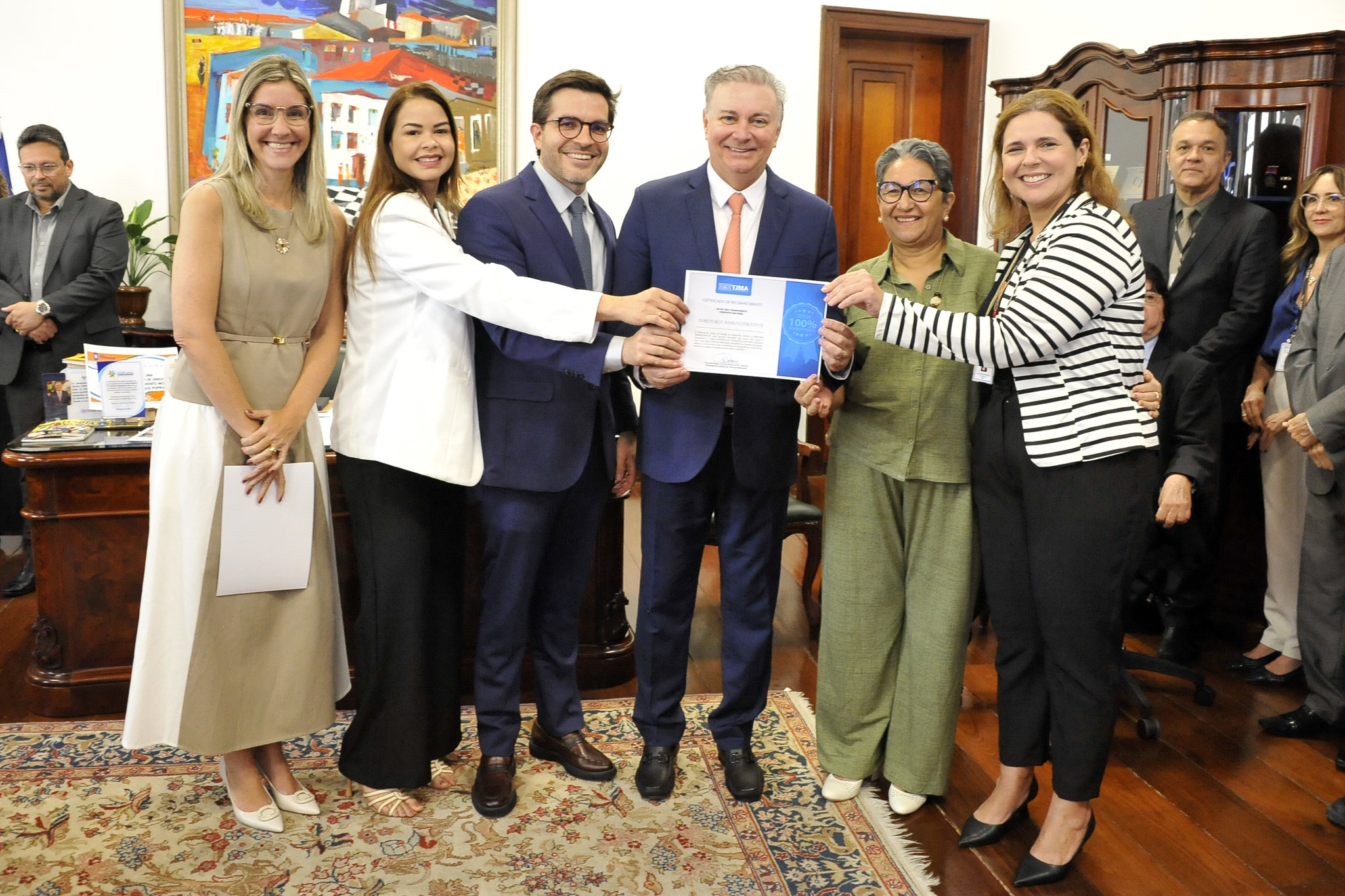 A imagem mostra um grupo de pessoas reunidas em um ambiente institucional elegante, com móveis de madeira, tapete decorativo e obras de arte coloridas nas paredes. Todos estão vestidos com roupas formais, reforçando o caráter oficial da ocasião. No centro da composição, duas pessoas seguram um certificado ¿ um gesto que indica a entrega de reconhecimento ou premiação.  O grupo está posicionado de forma organizada, sorrindo para a câmera, o que transmite um clima de celebração e valorização profissional. A ambientação e os gestos dos presentes sugerem que se trata de mais um registro da cerimônia de entrega dos certificados pelo desempenho no Ranking da Transparência, reforçando o espírito coletivo e o orgulho institucional.