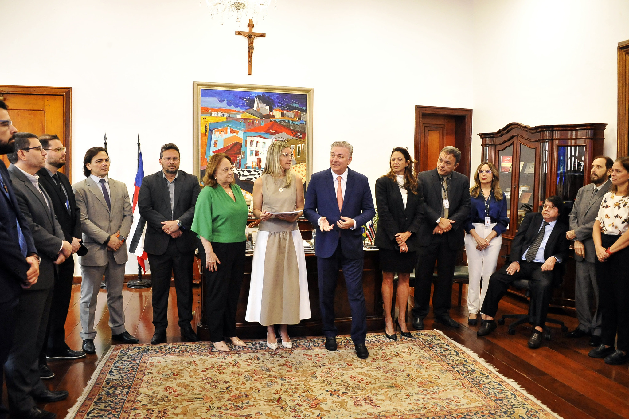 A imagem retrata uma solenidade em ambiente institucional refinado, com cerca de 17 pessoas reunidas em uma sala bem decorada. O espaço tem piso de madeira, tapete ornamental, móveis clássicos e uma pintura vibrante de paisagem urbana ao fundo, acima da qual há um crucifixo ¿ reforçando o caráter oficial e simbólico do local. Duas bandeiras posicionadas à esquerda indicam o contexto governamental ou judiciário.  O foco está em três pessoas ao centro: uma mulher com vestido bege segurando um documento, um homem de terno azul que parece estar discursando ou apresentando algo, e uma mulher de blusa verde. Os demais participantes, em trajes formais, estão dispostos em semicírculo, atentos e sorridentes, sugerindo um momento de reconhecimento ou anúncio institucional.