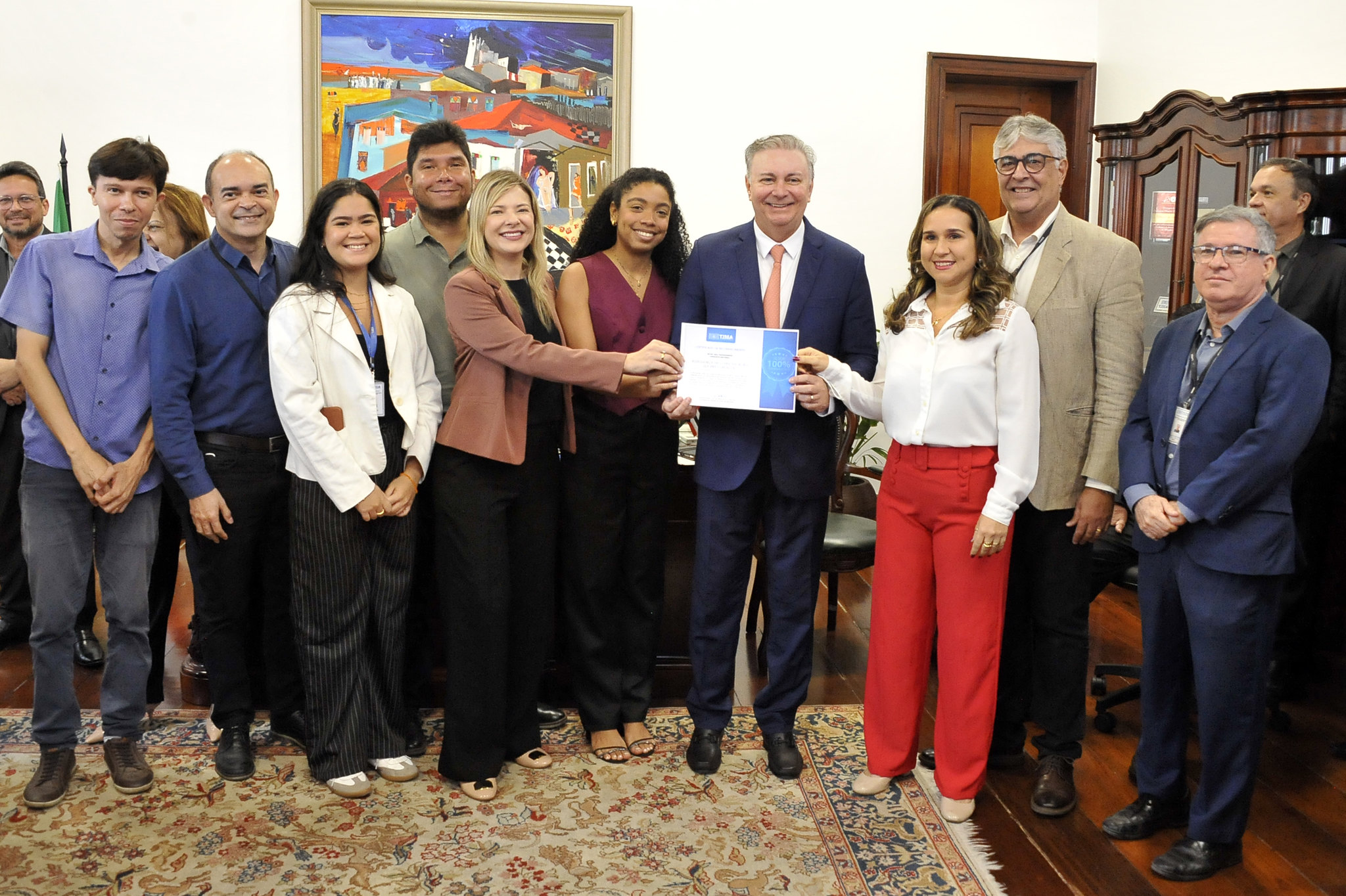 A imagem mostra um grupo de treze pessoas reunidas em um ambiente formal, provavelmente dentro de uma sede institucional ou gabinete. Todos estão vestidos com roupas sociais ou semi-formais, transmitindo profissionalismo e solenidade. No centro da composição, um homem de terno e gravata segura um certificado ao lado de uma mulher com blusa branca e calça vermelha, ambos sorrindo ¿ o que indica que estão sendo homenageados ou reconhecidos.  O grupo está posicionado em frente a uma parede decorada com uma pintura colorida, e há um móvel de madeira ao fundo, além de um tapete que reforça o tom elegante do espaço. A atmosfera é de celebração e reconhecimento, com todos os presentes demonstrando satisfação e orgulho pelo momento registrado.