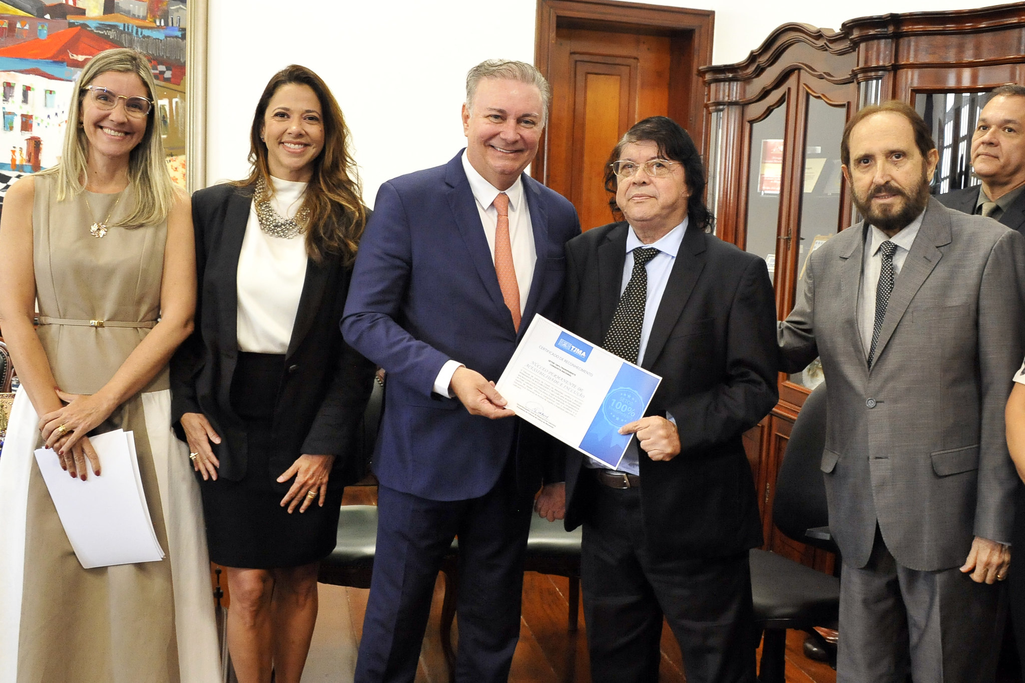 A imagem mostra um momento de entrega formal de certificado, capturado em ambiente institucional. No centro, um homem entrega um documento oficial a outro, ambos sorrindo, cercados por colegas em trajes sociais. O grupo está reunido em uma sala com móveis de madeira, armários ao fundo e quadros decorativos nas paredes, reforçando o clima de solenidade.  O certificado visível nas mãos dos homenageados indica que se trata de uma cerimônia de reconhecimento, provavelmente relacionada ao desempenho no Ranking da Transparência. A postura dos presentes e o registro fotográfico sugerem orgulho e celebração pelo feito alcançado.