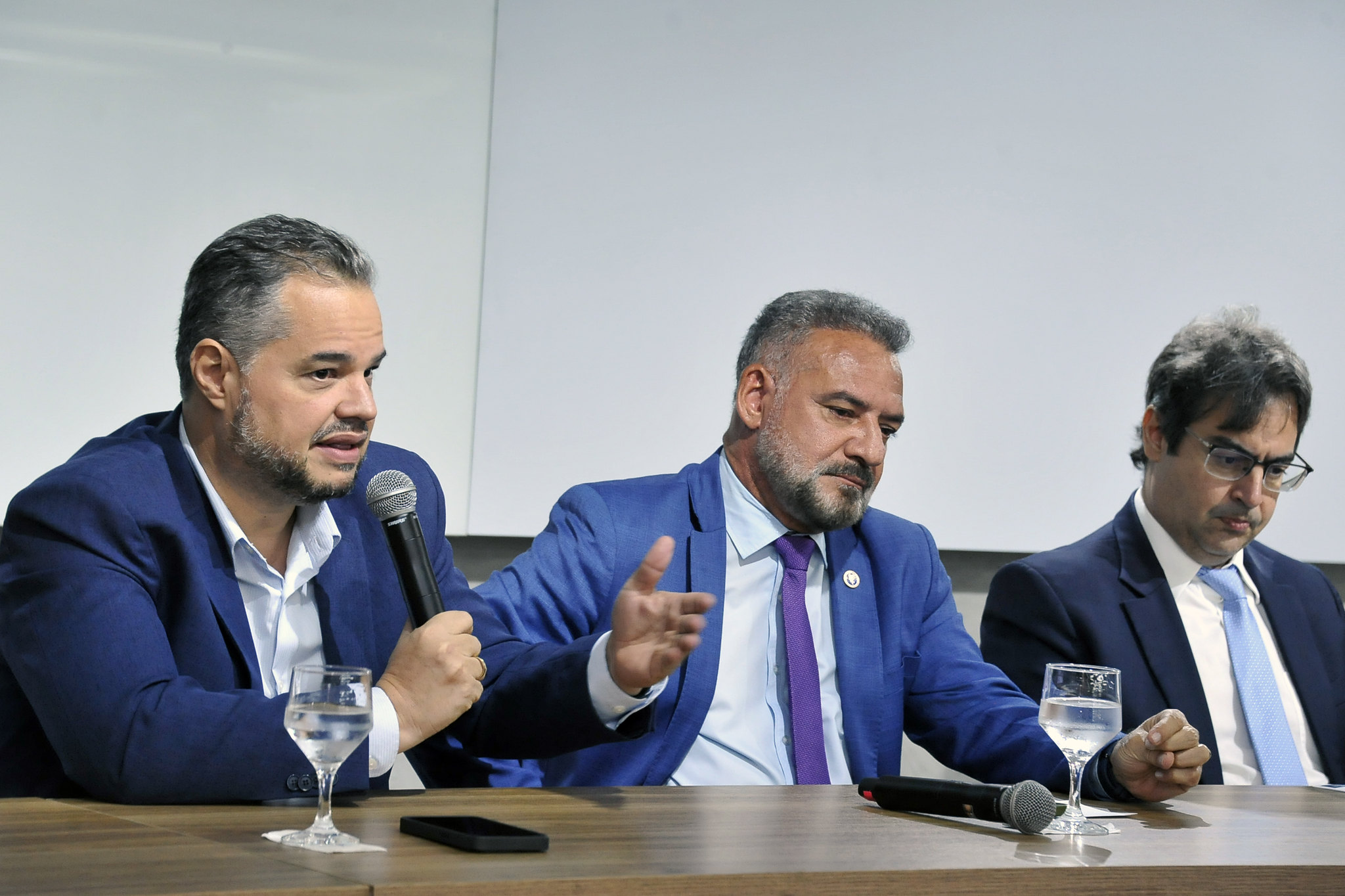 A imagem mostra uma mesa de painel com cinco pessoas sentadas, todas vestidas com roupas sociais, indicando um evento formal ¿ provavelmente uma conferência, mesa-redonda ou coletiva de imprensa. O homem ao centro está com um microfone na mão e parece estar falando, enquanto os outros escutam atentamente ou se preparam para contribuir.  Atrás deles há uma tela branca de projeção e, à direita, um banner com texto em português e um logotipo que remete à justiça ¿ uma figura estilizada segurando uma balança. O banner informa que o evento ocorre de 17 a 21 de setembro, na Sala Multiuso do Campus Renascer, o que reforça o caráter institucional e acadêmico da ocasião.  Tudo indica que o tema está relacionado ao direito ou à justiça, talvez uma semana jurídica ou um ciclo de debates sobre temas legais.