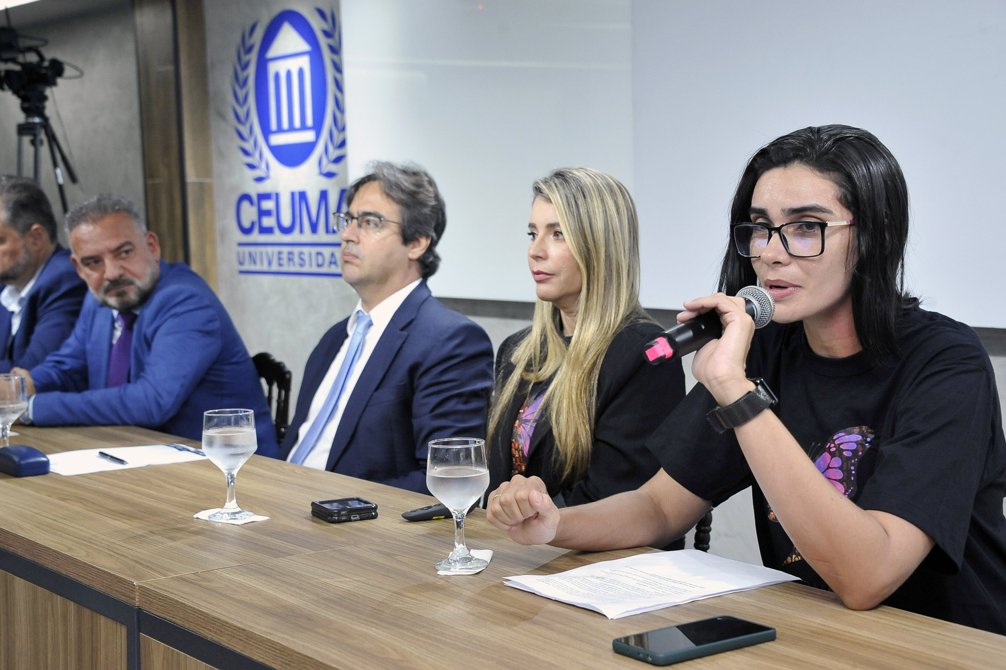 A imagem mostra uma mesa de painel com cinco pessoas sentadas, todas vestidas com roupas sociais, indicando um evento formal ¿ provavelmente uma conferência, mesa-redonda ou coletiva de imprensa. O homem ao centro está com um microfone na mão e parece estar falando, enquanto os outros escutam atentamente ou se preparam para contribuir.  Atrás deles há uma tela branca de projeção e, à direita, um banner com texto em português e um logotipo que remete à justiça ¿ uma figura estilizada segurando uma balança. O banner informa que o evento ocorre de 17 a 21 de setembro, na Sala Multiuso do Campus Renascer, o que reforça o caráter institucional e acadêmico da ocasião.  Tudo indica que o tema está relacionado ao direito ou à justiça, talvez uma semana jurídica ou um ciclo de debates sobre temas legais.