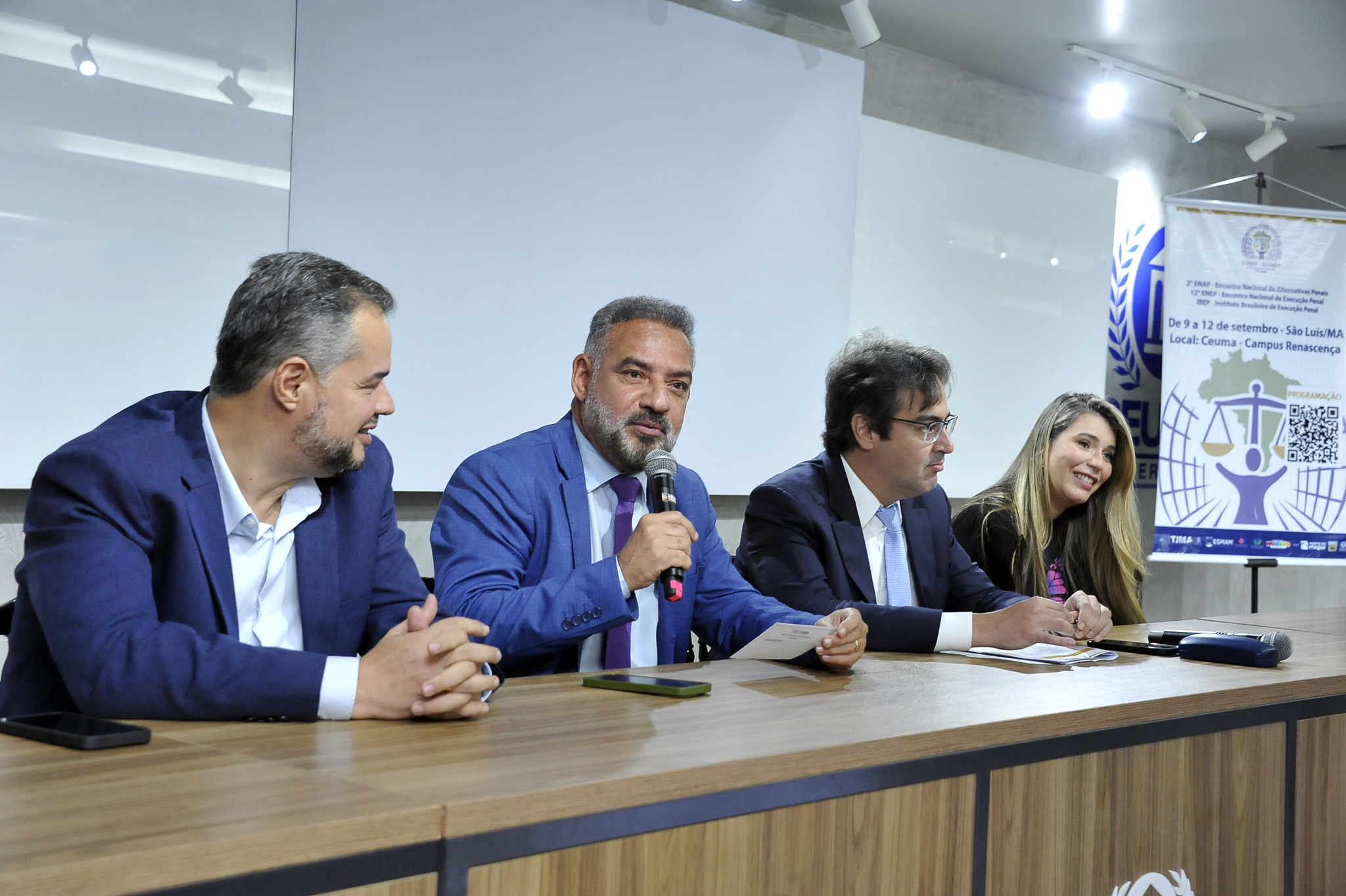 A imagem mostra uma mesa de painel com cinco pessoas sentadas, todas vestidas com roupas sociais, indicando um evento formal ¿ provavelmente uma conferência, mesa-redonda ou coletiva de imprensa. O homem ao centro está com um microfone na mão e parece estar falando, enquanto os outros escutam atentamente ou se preparam para contribuir.  Atrás deles há uma tela branca de projeção e, à direita, um banner com texto em português e um logotipo que remete à justiça ¿ uma figura estilizada segurando uma balança. O banner informa que o evento ocorre de 17 a 21 de setembro, na Sala Multiuso do Campus Renascer, o que reforça o caráter institucional e acadêmico da ocasião.  Tudo indica que o tema está relacionado ao direito ou à justiça, talvez uma semana jurídica ou um ciclo de debates sobre temas legais.