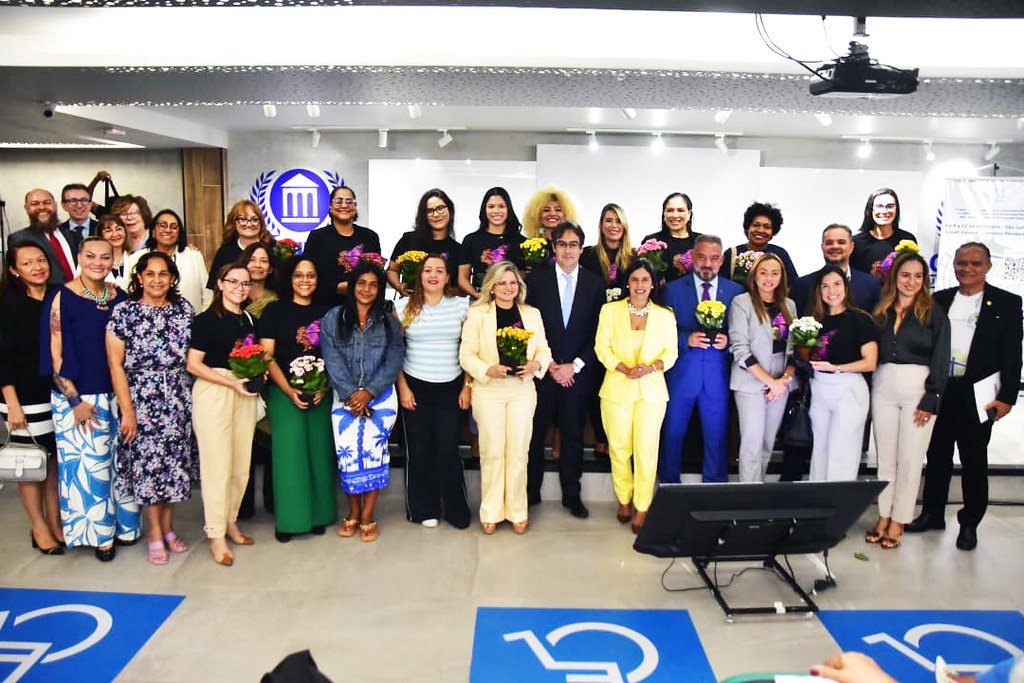 A imagem mostra uma mesa de painel com cinco pessoas sentadas, todas vestidas com roupas sociais, indicando um evento formal ¿ provavelmente uma conferência, mesa-redonda ou coletiva de imprensa. O homem ao centro está com um microfone na mão e parece estar falando, enquanto os outros escutam atentamente ou se preparam para contribuir.  Atrás deles há uma tela branca de projeção e, à direita, um banner com texto em português e um logotipo que remete à justiça ¿ uma figura estilizada segurando uma balança. O banner informa que o evento ocorre de 17 a 21 de setembro, na Sala Multiuso do Campus Renascer, o que reforça o caráter institucional e acadêmico da ocasião.  Tudo indica que o tema está relacionado ao direito ou à justiça, talvez uma semana jurídica ou um ciclo de debates sobre temas legais.