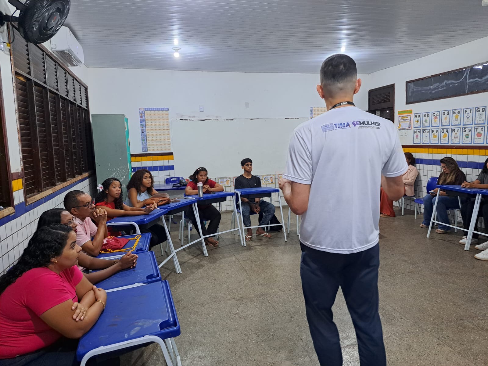 Em sala de aula, estudantes da Educação de Jovens e Adultos (EJA) estão sentados em carteiras azuis organizadas em formato de ¿U¿, atentos à fala de um integrante da equipe da CEMULHER/TJMA, que aparece em pé de costas para a câmera. O ambiente é simples, com paredes brancas, quadros didáticos fixados e janelas de madeira abertas.