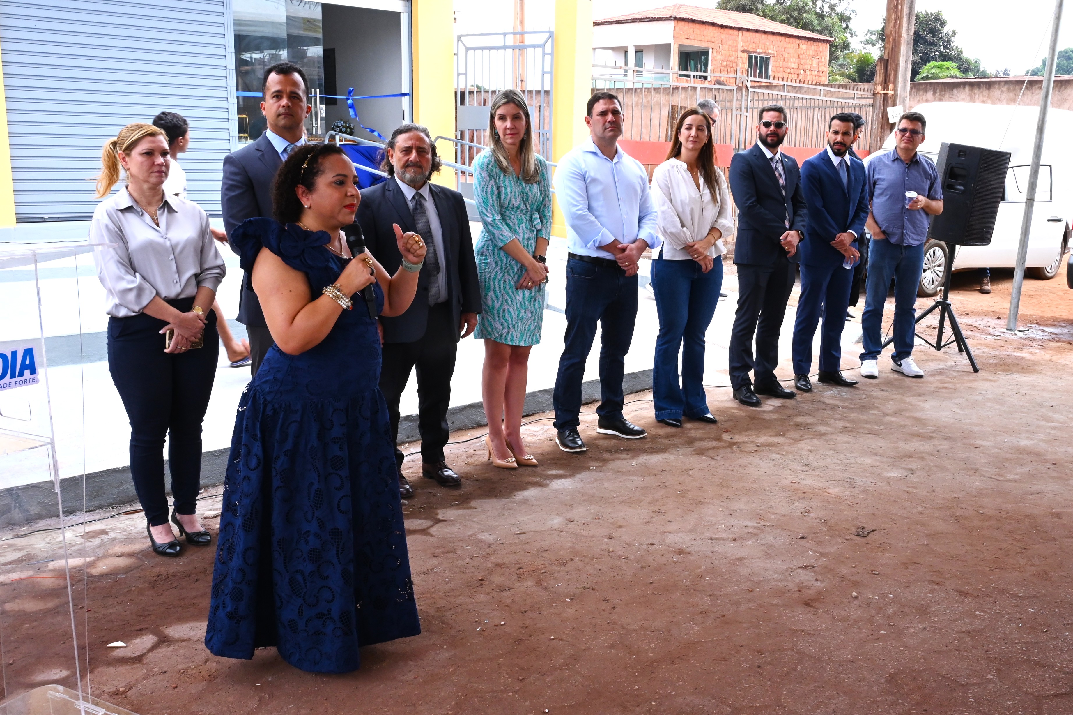Esta imagem é uma fotografia tirada ao ar livre que mostra um **momento de discurso** durante o evento de inauguração, com um grupo de autoridades ou convidados em segundo plano.  ---  ## Descrição da Imagem  ### Primeiro Plano (Oradora) No lado esquerdo, em primeiro plano, uma **mulher** com cabelos loiros e compridos está em pé, no que parece ser um **púlpito de acrílico transparente**.  * Ela está vestindo um **vestido curto estampado** em tons de verde-água e branco (o mesmo vestido da mulher sentada no escritório da foto DSC_9566.jpg). * Ela segura um **microfone** com a mão direita e gesticula com a esquerda, indicando que está **discursando** para a audiência (não visível na foto). * Abaixo do púlpito de acrílico, há um logotipo com a identificação da cidade: **AÇAILÂNDIA**.  ### Segundo Plano (Grupo de Autoridades) Atrás do púlpito, um **grupo de oito pessoas** está alinhado e posicionado em segundo plano. Eles parecem ser as autoridades e parceiros do evento.  * A maioria veste **trajes formais ou casuais de negócios**. Quatro homens estão de terno (cinza e azul-marinho). * A **mulher de vestido de festa azul-marinho** (também vista nas fotos anteriores) está no centro do grupo alinhado. * Há duas outras mulheres, uma de camisa social clara e calça escura, e outra de camisa azul clara e jeans. * Todos estão olhando em direção à oradora ou para frente, com uma postura formal.  ### Ambiente O cenário é de um **local externo e aberto**, com um piso de terra batida (cor marrom-avermelhada) em primeiro plano.  * **Fundo:** A fachada de um prédio está visível, com portas de vidro grandes e estruturas amarelas e azuis, possivelmente o mesmo prédio onde o Ponto de Inclusão Digital foi instalado. * **Detalhe:** No canto direito, parte de uma tenda ou lona e um **alto-falante preto** (caixa de som) confirmam que o local está preparado para um evento ao ar livre com som amplificado. Uma pessoa de camiseta preta e calça escura caminha no canto extremo direito.  A imagem registra o **momento de fala e apresentação** do projeto para o público.
