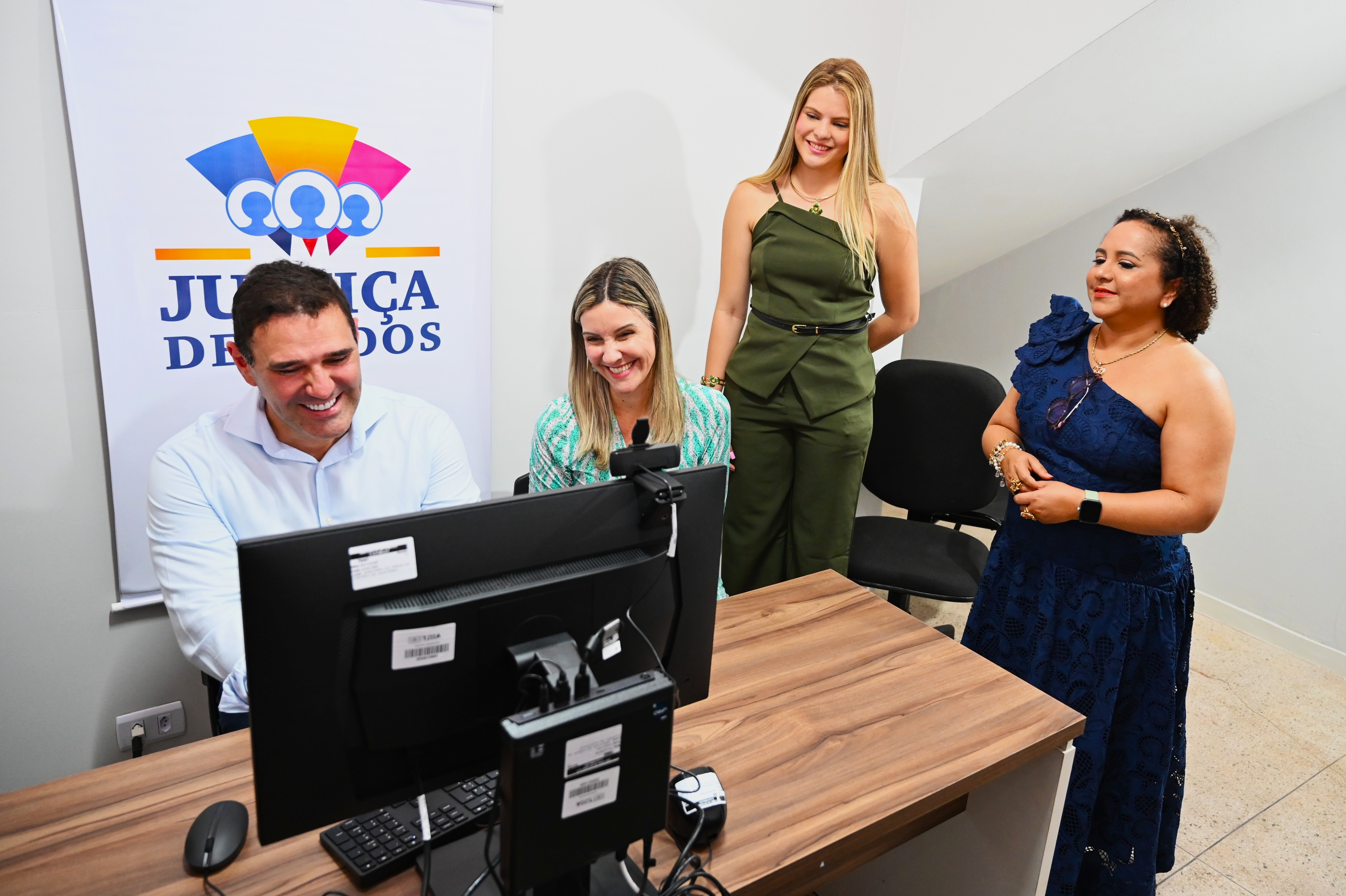 Esta imagem é uma fotografia de interior que mostra quatro pessoas em torno de um computador, em um ambiente que parece ser um escritório ou o recém-inaugurado **Ponto de Inclusão Digital** visto nas fotos anteriores.  ---  ## Descrição da Imagem  ### Pessoas e Interação Quatro indivíduos estão agrupados perto de uma mesa de trabalho, todos sorrindo e com expressões de satisfação.  1.  **Homem Sentado (à esquerda):** Um homem de camisa social azul clara (o mesmo da assinatura na foto anterior) está sentado na frente do monitor, olhando para a tela. Ele sorri de forma engajada. 2.  **Mulher Sentada (ao centro):** Uma mulher de cabelos loiros e compridos, vestindo uma blusa estampada em verde e branco, está sentada ao lado do homem, também olhando para a tela e sorrindo. Ela parece estar apontando ou interagindo com o conteúdo do monitor. 3.  **Mulher em Pé (atrás, ao centro):** Uma mulher loira, alta, vestindo um elegante conjunto de calça e *blazer* sem manga verde-oliva, está em pé atrás da mesa, observando a tela com interesse e um sorriso. 4.  **Mulher em Pé (à direita):** A mulher de vestido de festa azul-marinho de um ombro só, também vista nas fotos anteriores, está em pé ao lado da mesa, olhando para o grupo.  A **interação** sugere que eles estão revisando ou demonstrando o funcionamento de um sistema ou serviço digital no local.  ### Ambiente e Contexto O local é um **escritório moderno e limpo**:  * **Mobiliário:** Eles estão em volta de uma **mesa de trabalho** de madeira escura, com um monitor de computador, teclado e mouse. * **Fundo:** A parede do fundo é branca. * **Identificação:** No canto superior esquerdo, há um **banner** ou cartaz vertical pendurado na parede, com um grande **logotipo colorido** e a frase **"JUSTIÇA DE TODOS"** em destaque. * **Cadeiras:** Duas cadeiras de escritório pretas estão vazias no lado direito, atrás da mulher de verde-oliva.  O cenário interno confirma o propósito do local: um ambiente estruturado para o acesso a **serviços digitais de justiça ou cidadania**, como parte do projeto "Justiça de Todos" e o "Ponto de Inclusão Digital".