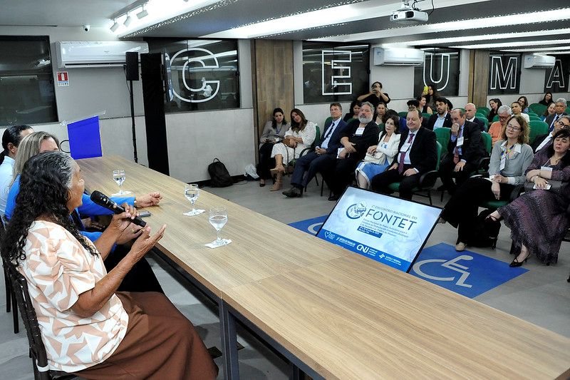 Foto horizontal de encontro do Fontet em São Luís. A imagem mostra uma reunião ou evento em ambiente interno. Na parte frontal e à esquerda, há uma mesa de conferência comprida e de madeira. Sentadas atrás dela estão algumas pessoas, sendo que uma mulher idosa de cabelos longos e grisalhos, com uma blusa de estampa clara, está falando ao microfone. Do outro lado da mesa, de frente para a audiência, há cadeiras ocupadas por diversas pessoas em trajes formais. A plateia está atenta ao que está sendo dito. No canto inferior direito, há uma tela de monitor com o logotipo do "VI ENCONTRO NACIONAL DO FONTET" e os logotipos do "CNJ" e da "Justiça Federal". As paredes do local são revestidas de madeira e há letras grandes e soltas formando a sigla "TJMA" em uma das paredes ao fundo.