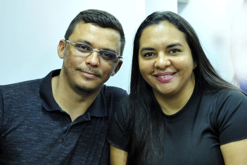 Foto horizontal: Mulher oficializa acordo e união estável na Conciliação do TJMA. A foto mostra um casal sorrindo e olhando para a câmera. O homem está do lado esquerdo da foto, usando óculos e uma camisa polo escura, enquanto a mulher está do lado direito, com cabelos longos e escuros e usando uma blusa preta. Ambos têm expressões felizes e descontraídas.