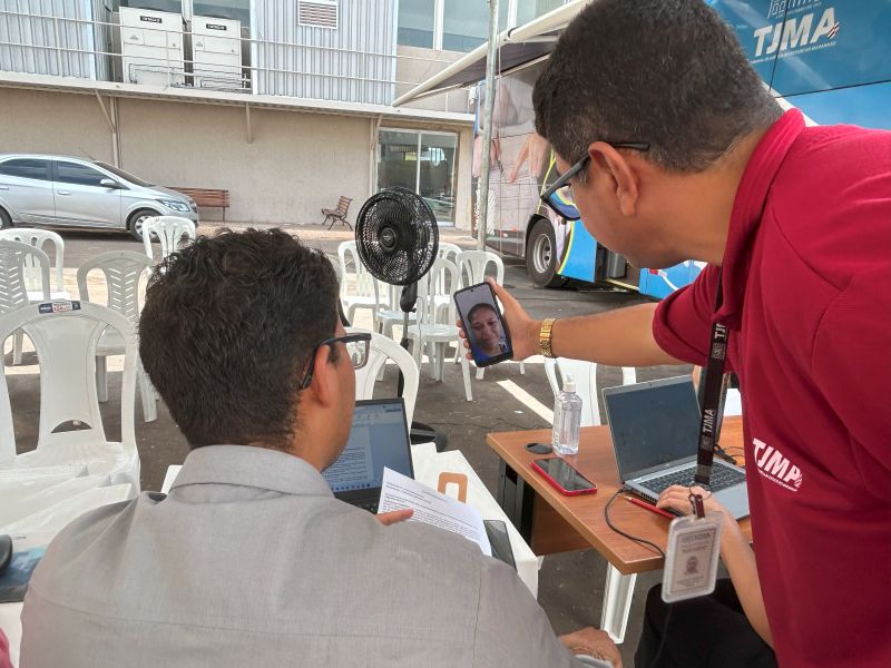Foto horizontal: Mulher oficializa acordo e união estável na Conciliação do TJMA. A foto mostra duas pessoas em uma mesa de trabalho montada ao ar livre, em frente a um ônibus. Um está de costas para a câmera, com uma camisa cinza clara, olhando para um celular. O outro, que está de lado, usa uma camisa polo vermelha, e segura um celular com uma mão e um documento com a outra, mostrando a tela do celular para o primeiro homem.
