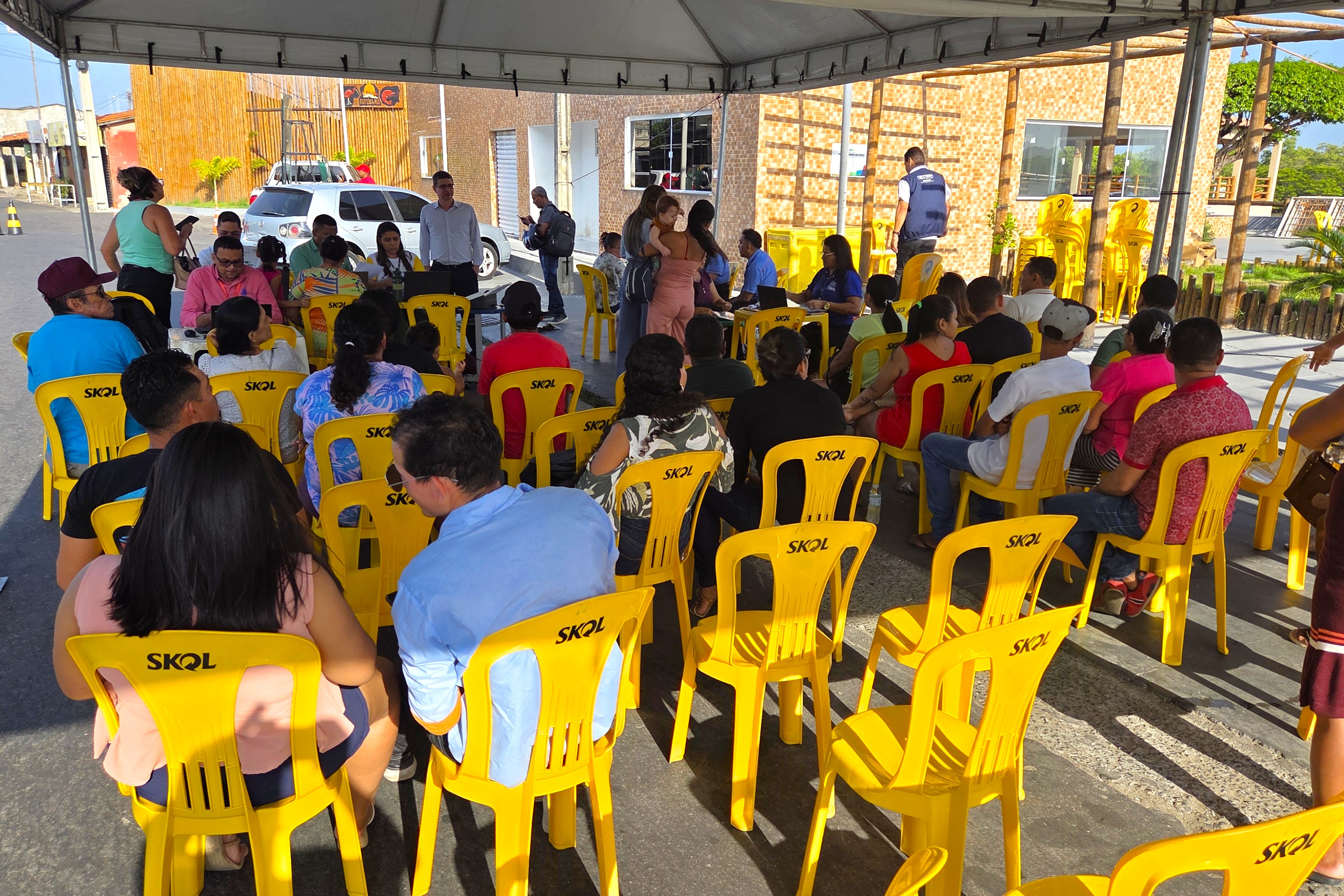 A imagem mostra uma reunião ou evento ao ar livre, sob uma tenda branca para proteção do sol. O público está sentado de costas para a câmera, em fileiras de cadeiras amarelas de plástico, a maioria com o logo da marca Skol.  Há um grupo de pessoas em pé no fundo, algumas parecem ser organizadores ou anfitriões do evento. Uma mulher de blusa verde está do lado esquerdo, e um homem de camisa azul, no centro, interage com o público.  O ambiente parece ser em uma área urbana, com a calçada e o asfalto visíveis, além de uma construção com paredes de tijolos e uma casa ao fundo. A iluminação é natural e o céu está claro. A cena sugere um evento comunitário, talvez uma palestra ou uma assembleia.