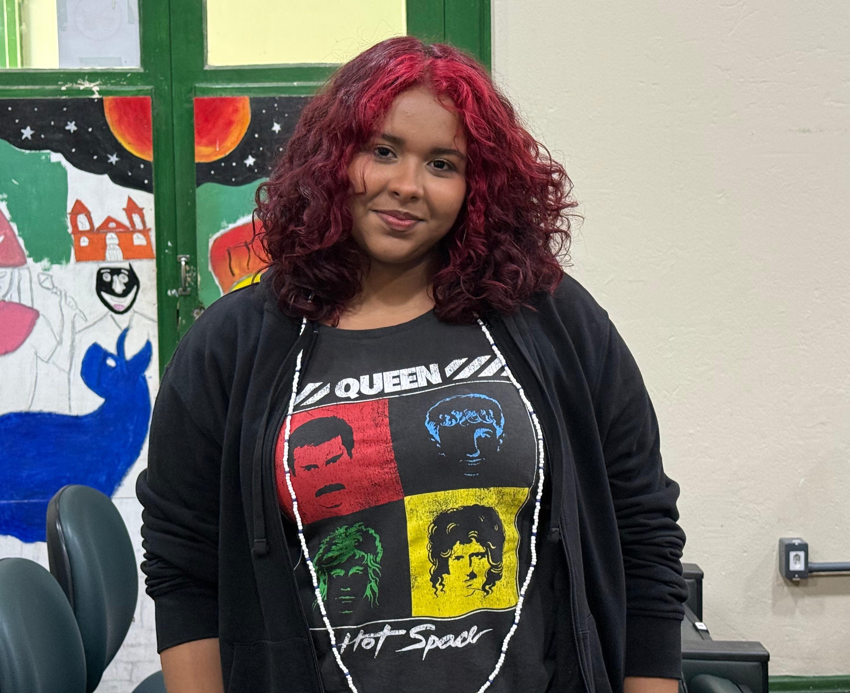 A estudante Sâmarah Emilly Pinheiro, do primeiro período do curso de Artes Visuais do IFMA, aparece em destaque em frente a um mural colorido com desenhos artísticos. Ela veste camiseta preta da banda Queen e casaco aberto, e tem os cabelos cacheados tingidos de vermelho.