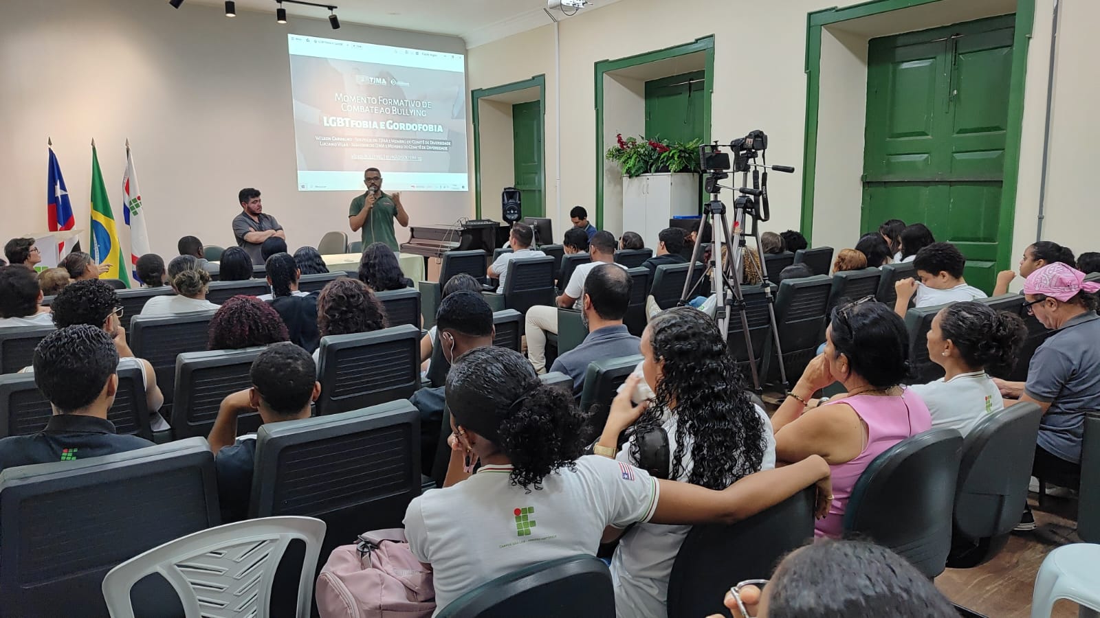 Auditório do IFMA Centro Histórico com estudantes sentados em cadeiras enfileiradas, atentos à fala dos servidores do TJMA, Luciano Vilar e Wilson Carvalho, integrantes do Comitê de Diversidade. Ao fundo, a tela projeta o tema da palestra, enquanto câmeras registram a atividade.