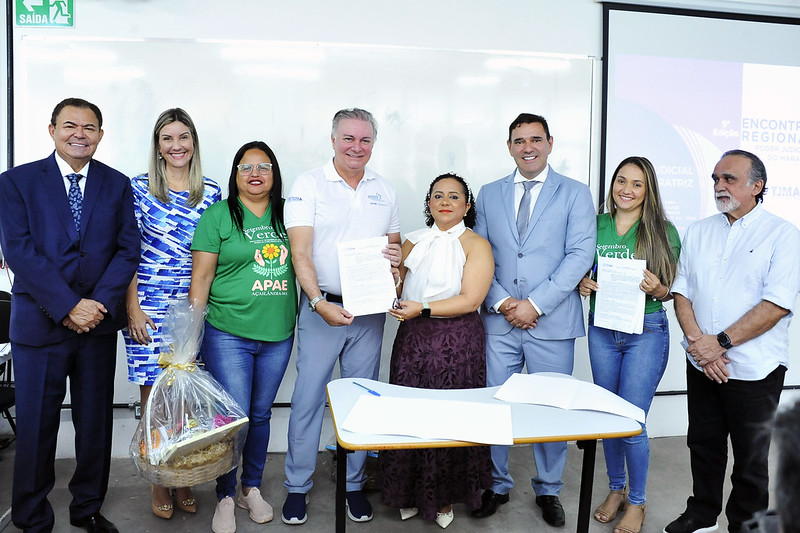 Foto horizontal de evento em que o TJMA assina termo com Apae de Açailândia e doa veículos a dois municípios. Nesta imagem, sete pessoas estão posando para uma foto em um ambiente interno. No centro da imagem, um homem de terno azul claro e uma mulher com uma blusa branca de laço estão segurando um documento. Ao lado esquerdo deles, uma mulher de camiseta verde com a inscrição "Orgulho Verde APAE Açailândia" e um homem de terno azul escuro. A mulher da camiseta segura uma cesta de presente. Do lado direito do casal central, há um homem de terno cinza, uma mulher de camiseta verde e um homem de barba de terno branco.