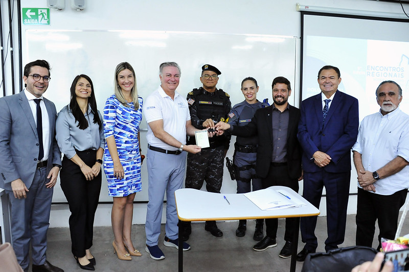Foto horizontal de evento em que o TJMA assina termo com Apae de Açailândia e doa veículos a dois municípios. A imagem mostra um evento oficial onde um grupo de pessoas está posando para uma foto. No centro, um homem de camisa polo branca está entregando chaves a um oficial de polícia militar. As pessoas na imagem estão vestidas de forma profissional e formal, indicando a importância do evento. Ao fundo, um quadro branco com a palavra "SAÍDA" e um pôster com o texto "ENCONTROS REGIONAIS" também são visíveis.