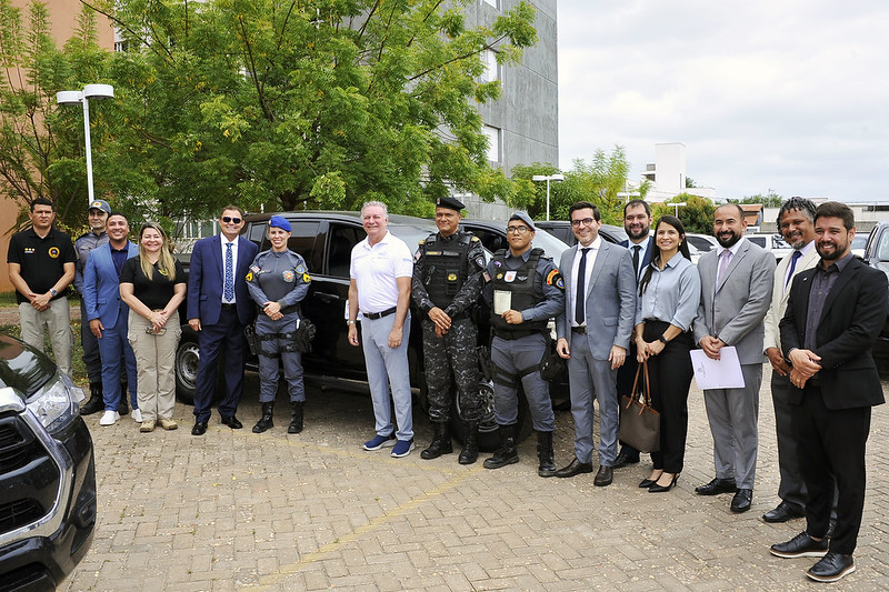 Foto horizontal de evento em que o TJMA assina termo com Apae de Açailândia e doa veículos a dois municípios. A imagem mostra um grupo de pessoas em frente a veículos estacionados. A maioria das pessoas está vestindo roupas formais, como ternos e camisas sociais, mas há também alguns indivíduos com uniformes militares, incluindo uma mulher e um homem com óculos escuros.