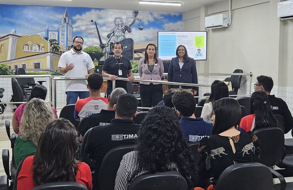 A foto mostra quatro pessoas de pé na frente da sala, de frente para o público. Da esquerda para a direita, há um homem com óculos e barba, vestindo uma polo branca, um homem com camisa preta, uma mulher com um blazer rosa claro e uma mulher com um blazer azul escuro. Todos estão sorrindo e olhando para a plateia. A sala tem uma parede de fundo com um mural pintado, que retrata uma igreja, uma figura da justiça com uma balança e uma paisagem urbana. Um monitor na parede mostra um slide com texto e uma imagem. O público está sentado de costas para a câmera, olhando para os palestrantes. Alguns usam camisetas pretas com o logo "TJMA".
