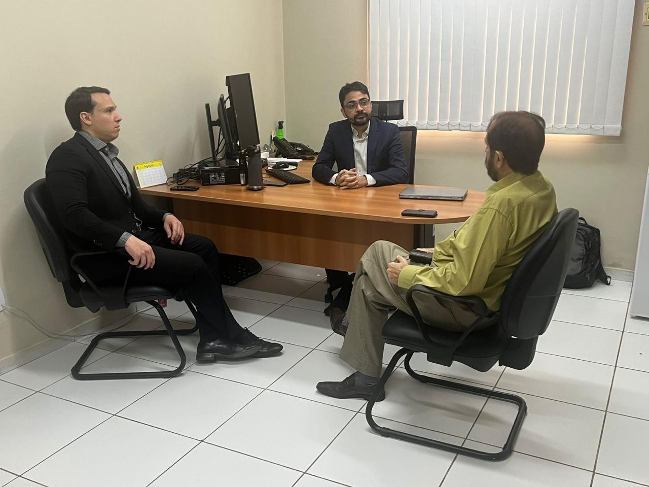 Em uma sala de escritório, três homens estão sentados, conversando em torno de uma mesa de madeira. À esquerda, um homem de terno preto e camisa cinza observa atentamente. Ao centro, atrás da mesa, um homem de terno azul-marinho e camisa branca fala, com um notebook e um celular sobre a mesa. À direita, outro homem, com camisa social verde clara e calça bege, ouve a conversa. Ao fundo, há uma janela com persiana branca e um computador sobre a mesa.