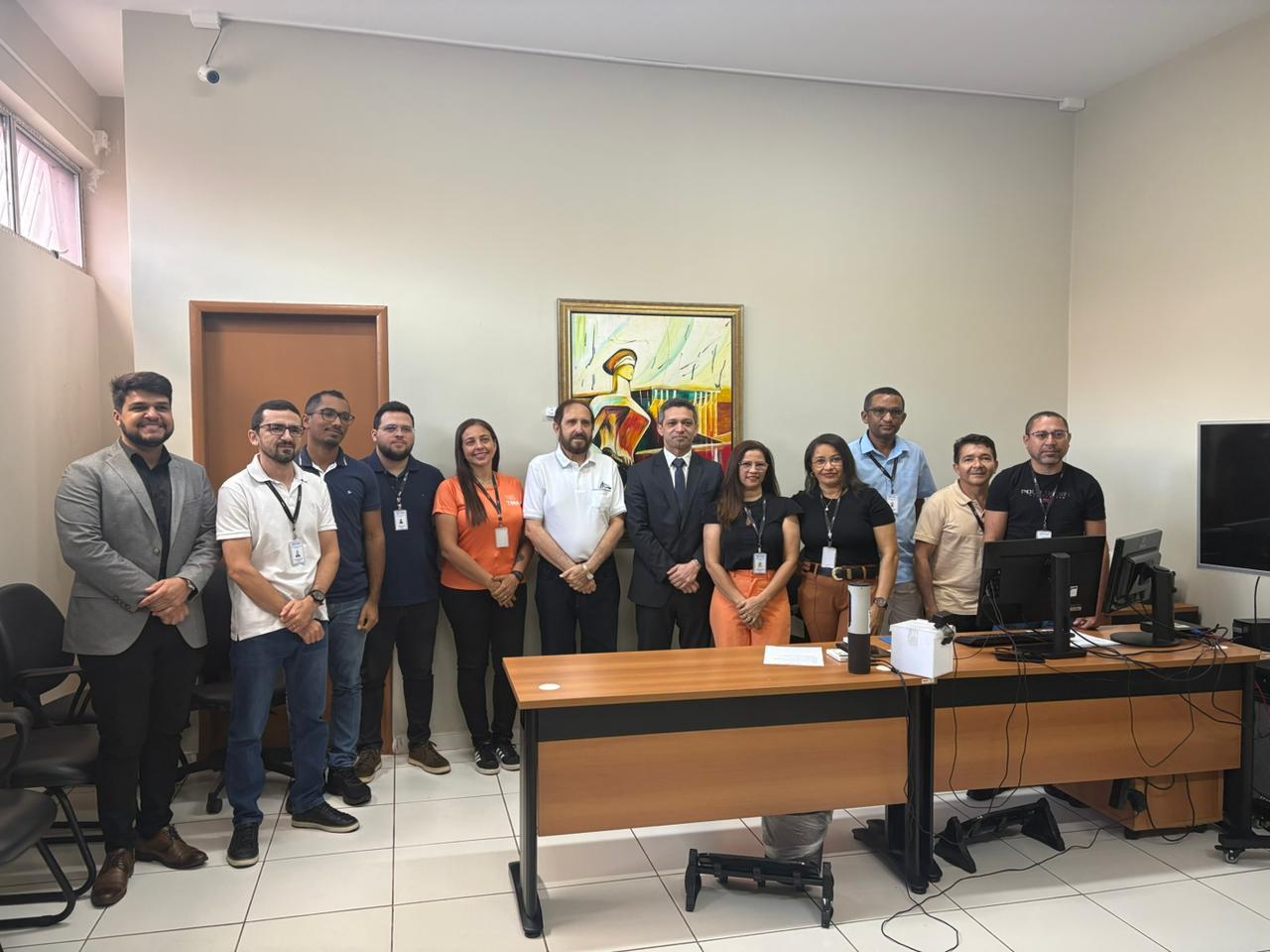 Quinze pessoas, entre homens e mulheres, estão em pé, lado a lado, em uma sala de audiências. À esquerda e à direita, há mesas com computadores, e ao fundo, uma parede clara com um quadro colorido pendurado. Alguns participantes usam roupas formais, outros estão com vestimenta mais casual, e vários têm crachás do TJMA. Todos olham para a câmera e sorriem.