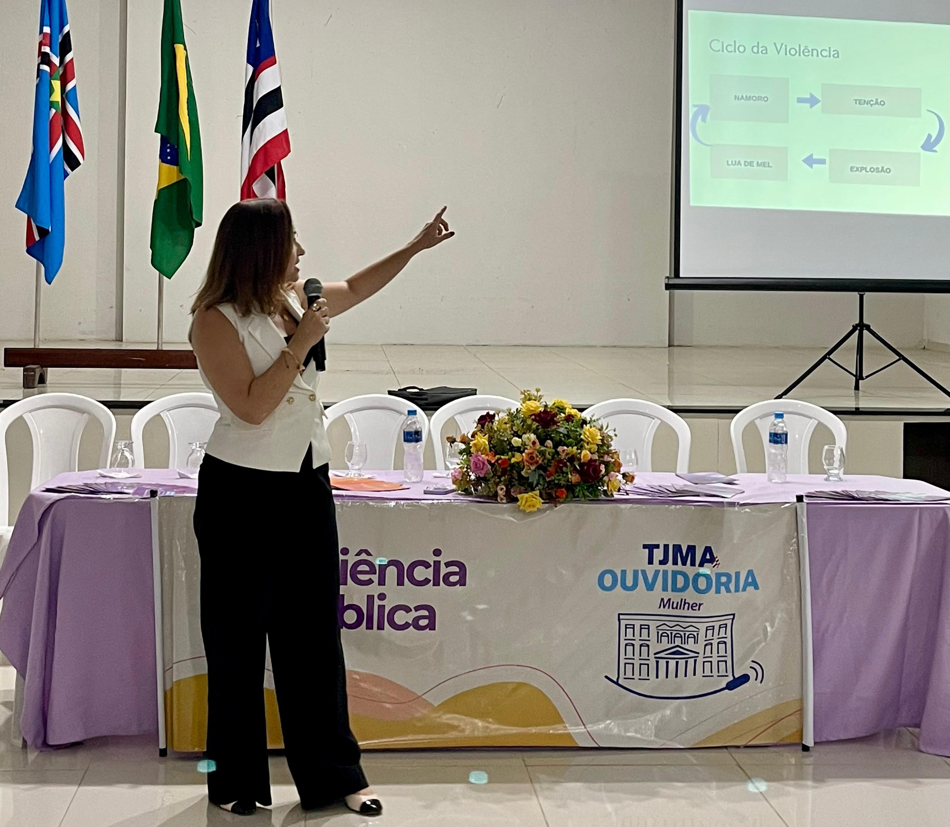 Mulher em pé, à frente da mesa com toalha lilás e arranjo floral, segura um microfone e aponta para um slide projetado na tela. A apresentação exibe o título ¿Ciclo da Violência¿ e um fluxograma com setas ligando palavras como ¿Namoro¿, ¿Tensão¿, ¿Explosão¿ e ¿Lua de Mel¿. As bandeiras do Maranhão, do Brasil e outra estadual/municipal estão posicionadas no palco ao fundo.