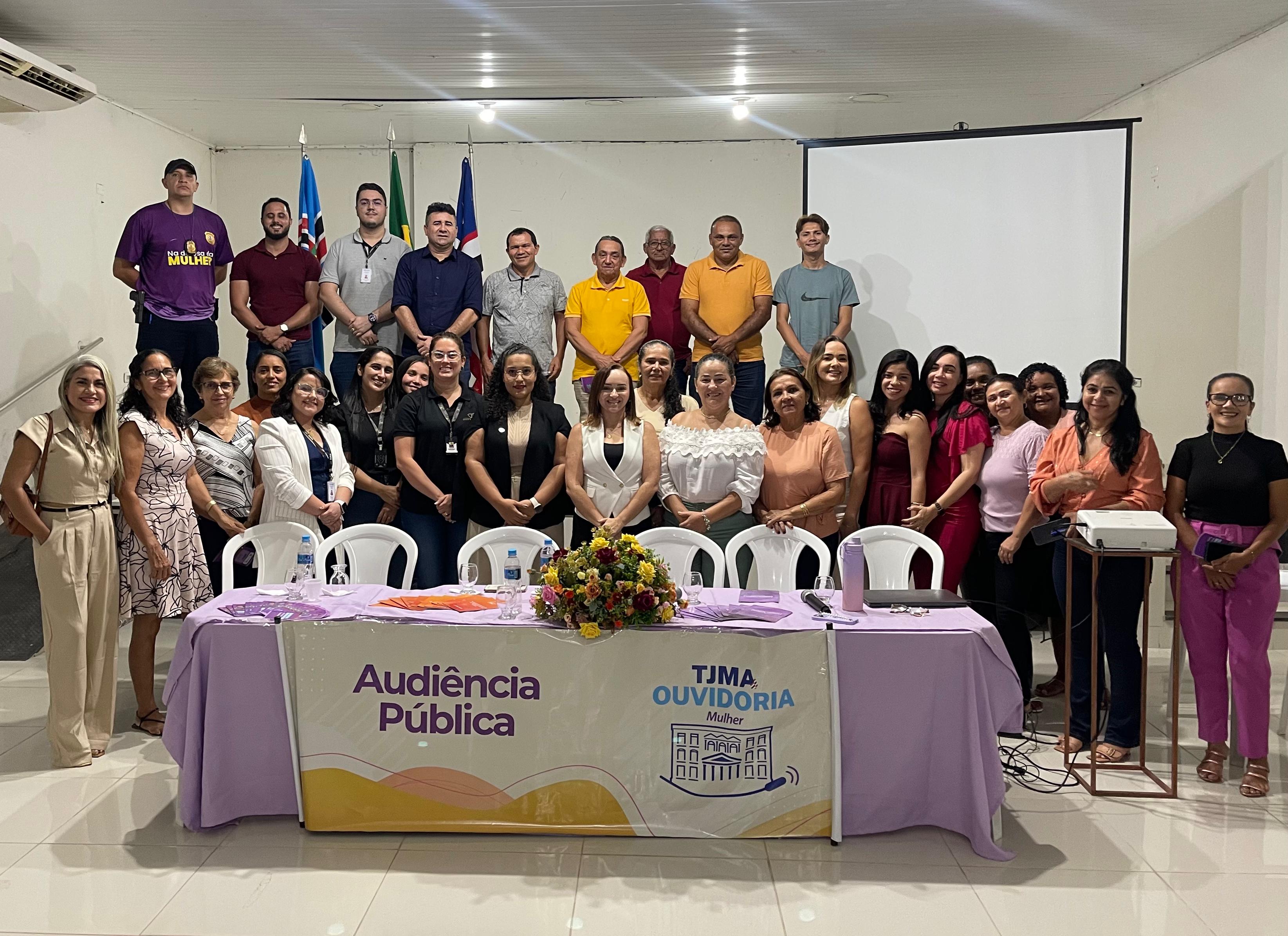 Grupo diverso de cerca de 30 pessoas posando para foto em um auditório. À frente, há uma mesa coberta com toalhas lilás e branca, com arranjo de flores coloridas ao centro e faixa com os dizeres ¿Audiência Pública¿ e o logotipo da Ouvidoria da Mulher do TJMA. Ao fundo, três bandeiras (do Maranhão, do Brasil e outra estadual/municipal) estão posicionadas na parede. As pessoas estão dispostas em duas fileiras, sorrindo para a câmera.