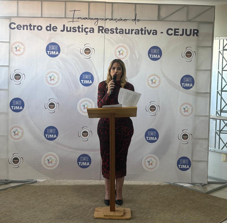 Juíza Larrisa Tupinambá fala ao microfone em um púlpito de madeira, segurando papéis em uma das mãos. Ao fundo, há um painel institucional com a identificação ¿Centro de Justiça Restaurativa ¿ CEJUR¿ e as logomarcas do TJMA e da FAESF.