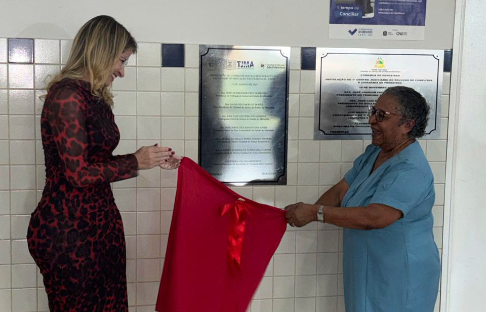 Duas mulheres realizam o descerramento de uma placa de inauguração afixada na parede. Uma delas segura um tecido vermelho, enquanto a outra, ao lado, acompanha sorridente o momento da revelação da placa.