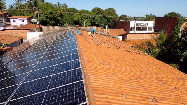 Imagem colorida. Foto de trabalhadores concluindo montagem de placas de energia solar em telhado de unidade judicial da Raposa.