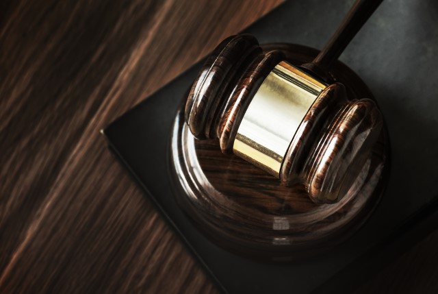 Fotografia colorida mostra um martelo de sentença de juiz, na cor marrom, sobre apoio da mesma cor.