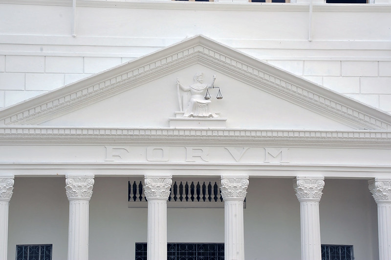 Foto horizontal de parte da fachada do TJMA. A imagem mostra a fachada do edifício com arquitetura clássica. A fachada possui colunas coríntias e um frontão triangular. No centro do frontão, há uma escultura de uma figura feminina segurando uma balança, simbolizando a deusa da justiça, Themis. Abaixo do frontão, está a palavra "FORVM" esculpida em letras maiúsculas.