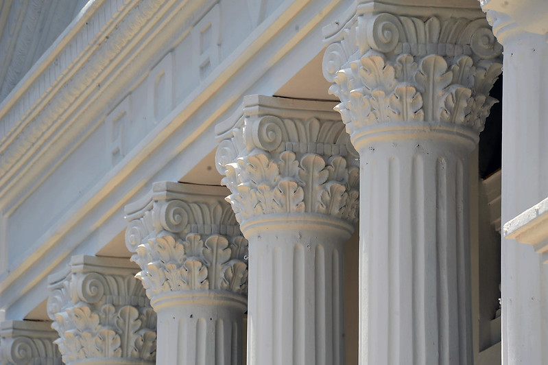 Foto horizontal de parte da fachada do TJMA. A imagem mostra uma série de colunas coríntias, um estilo de coluna arquitetônica caracterizado por capitéis ornamentados com folhas de acanto e volutas. As colunas são feitas de um material claro, possivelmente mármore, e estão alinhadas em uma estrutura do edifício clássico. A parte superior das colunas, os capitéis, é ricamente decorada com detalhes esculpidos, incluindo folhas e espirais. A imagem destaca a simetria e a elegância das colunas, que são elementos arquitetônicos importantes em muitos edifícios históricos e monumentos.