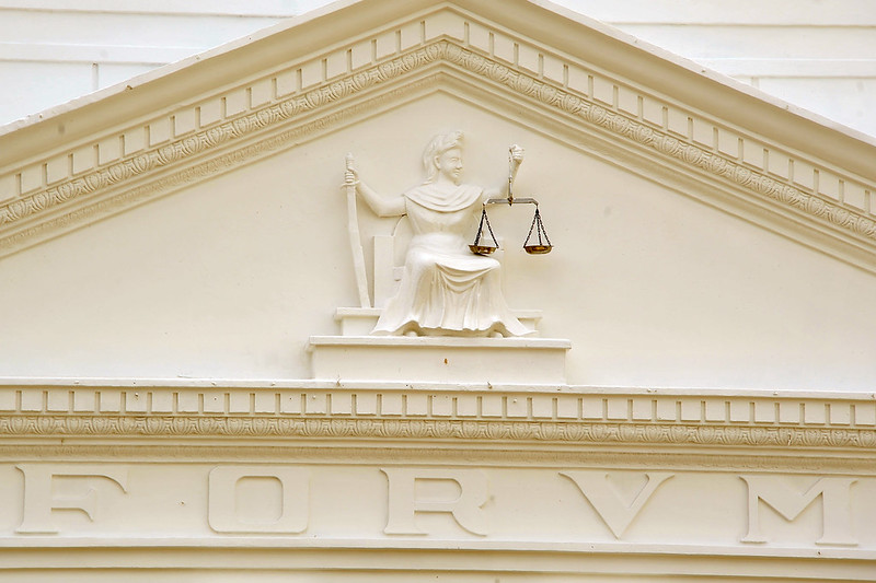 Foto horizontal de parte da fachada do TJMA. A imagem mostra um relevo no edifício do Tribunal. No centro do relevo, há uma figura feminina sentada, segurando uma balança na mão esquerda e uma espada na mão direita, que representa a deusa da Justiça, Themis. A balança é um símbolo clássico da justiça, representando a imparcialidade e o equilíbrio. A espada simboliza a autoridade e a capacidade de impor decisões. A figura está vestida com uma toga, que é tradicionalmente associada à justiça e à lei. Abaixo da figura, há letras esculpidas que formam a palavra "FORVM".