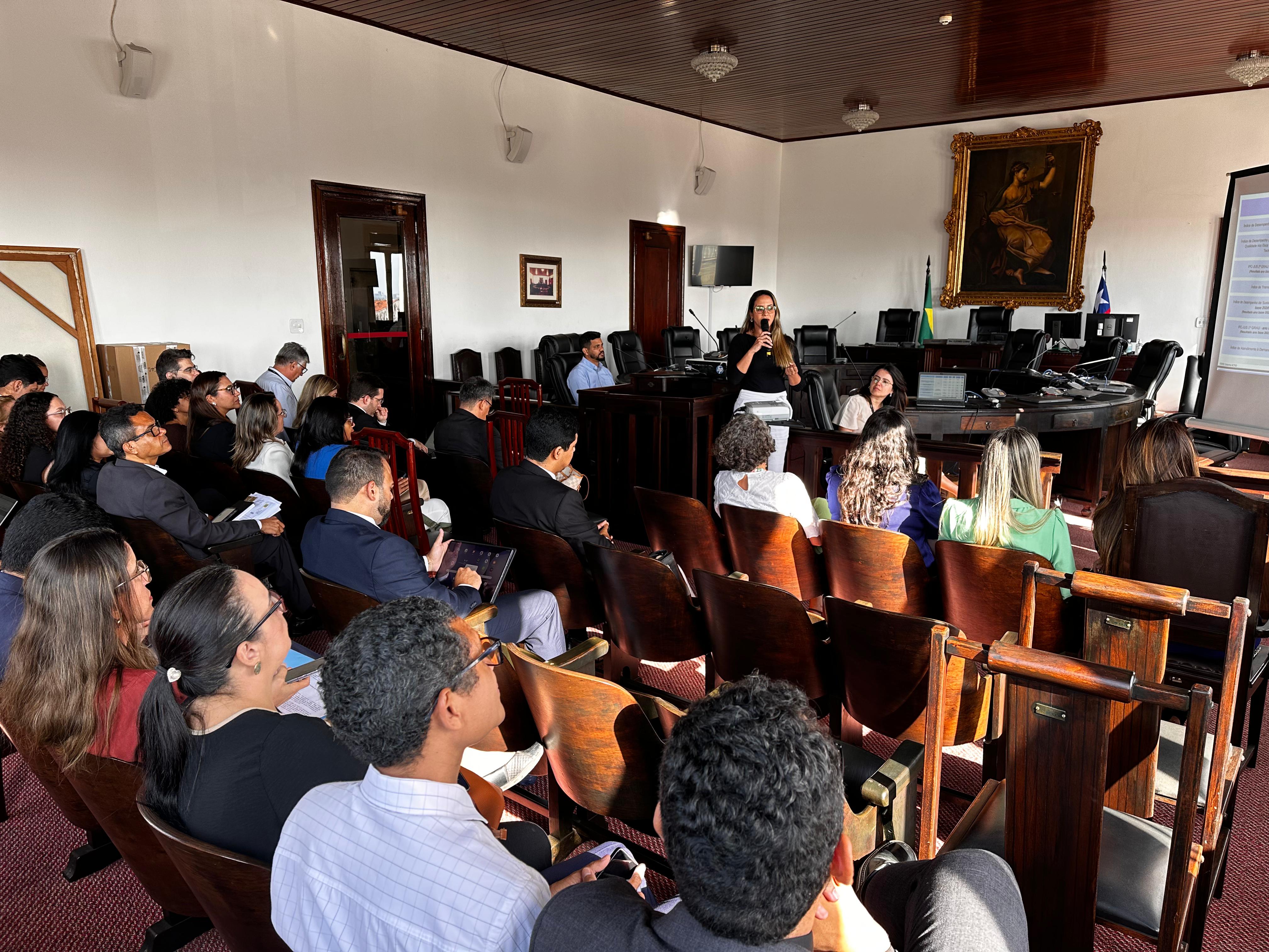 foto de sala ampla com um carpete vermelho sob seus pés. À sua esquerda, você sente várias fileiras de cadeiras de madeira escura, a maioria delas ocupada por pessoas atentas. Elas parecem estar voltadas para a frente da sala.À sua direita, há uma área ligeiramente elevada, onde você pode sentir uma mesa grande e robusta, também de madeira escura. Uma mulher está em pé atrás dessa mesa, segurando um microfone na mão direita e gesticulando com a esquerda enquanto fala. Ela parece estar conduzindo uma apresentação ou palestra.Você pode sentir a presença de um projetor posicionado em algum lugar à frente, provavelmente exibindo informações em uma tela que você não pode ver. Há também alguns equipamentos eletrônicos sobre a mesa elevada, talvez um computador e outros dispositivos.As paredes da sala parecem claras, e você pode sentir a presença de algumas portas e talvez janelas, permitindo a entrada de alguma luz. A atmosfera geral sugere um ambiente formal, como uma sala de reuniões ou um auditório pequeno, onde uma apresentação está em andamento para um público atento.