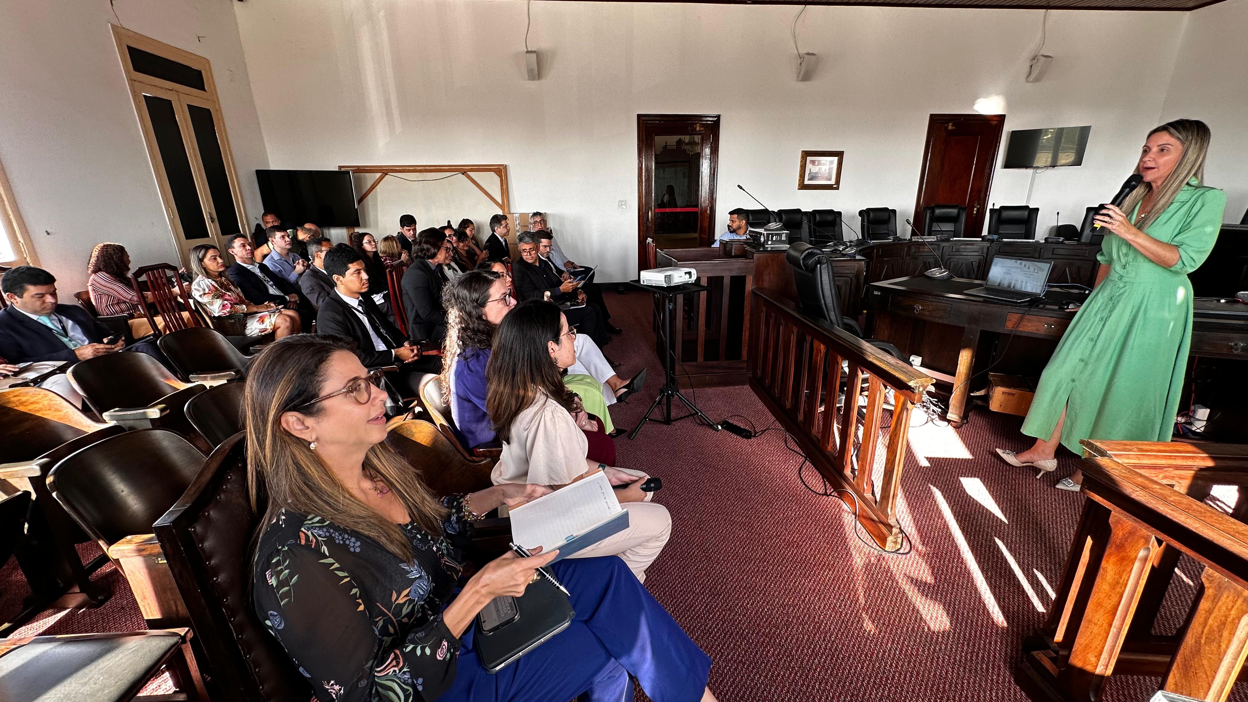 foto de sala ampla com um carpete vermelho sob seus pés. À sua esquerda, você sente várias fileiras de cadeiras de madeira escura, a maioria delas ocupada por pessoas atentas. Elas parecem estar voltadas para a frente da sala.À sua direita, há uma área ligeiramente elevada, onde você pode sentir uma mesa grande e robusta, também de madeira escura. Uma mulher está em pé atrás dessa mesa, segurando um microfone na mão direita e gesticulando com a esquerda enquanto fala. Ela parece estar conduzindo uma apresentação ou palestra.Você pode sentir a presença de um projetor posicionado em algum lugar à frente, provavelmente exibindo informações em uma tela que você não pode ver. Há também alguns equipamentos eletrônicos sobre a mesa elevada, talvez um computador e outros dispositivos.As paredes da sala parecem claras, e você pode sentir a presença de algumas portas e talvez janelas, permitindo a entrada de alguma luz. A atmosfera geral sugere um ambiente formal, como uma sala de reuniões ou um auditório pequeno, onde uma apresentação está em andamento para um público atento.
